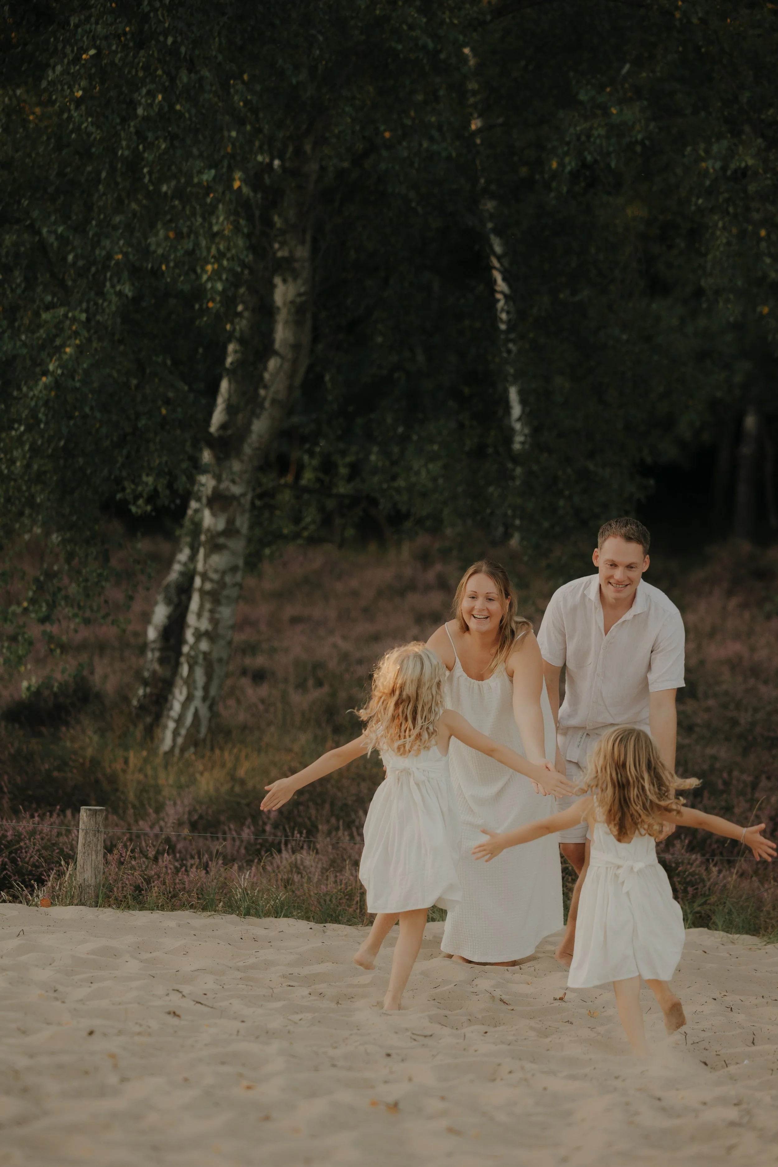 Familie, bestehend aus zwei Erwachsenen und zwei Kindern, läuft lachend und spielend im Sand im Freien, umgeben von Bäumen.
