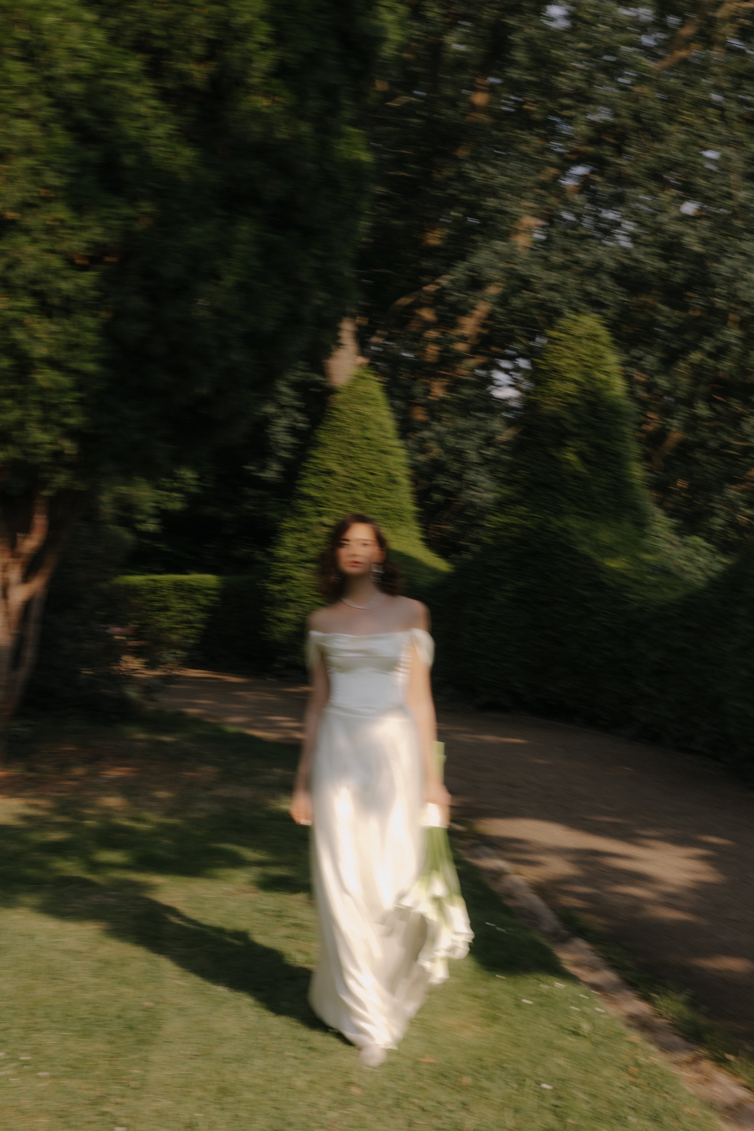Eine junge Frau in einem weißen Brautkleid läuft durch einen Garten mit topiary-Bäumen.