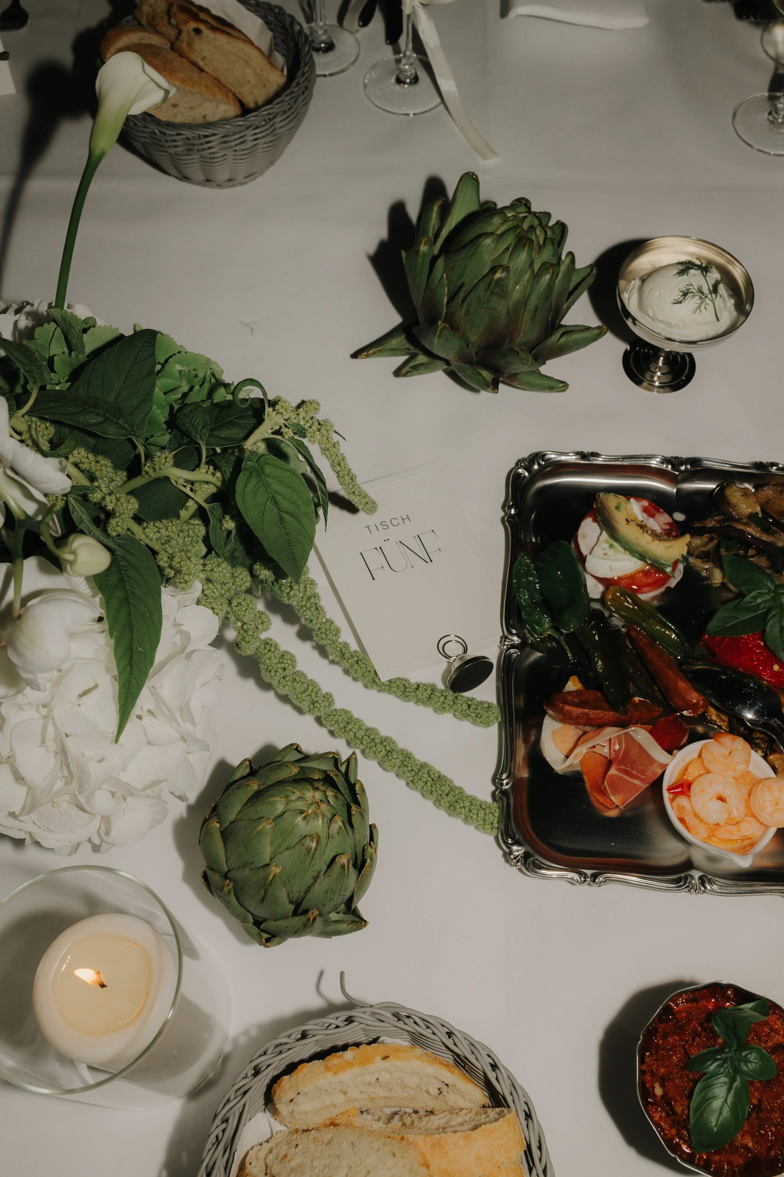 Ein gedeckter Tisch mit Blumen, Artischocken, einer Kerze, Brot, einer Platte mit Meeresfrüchten und Antipasti, sowie Gläsern und einem Schild mit der Aufschrift 'Tisch Fünf'.