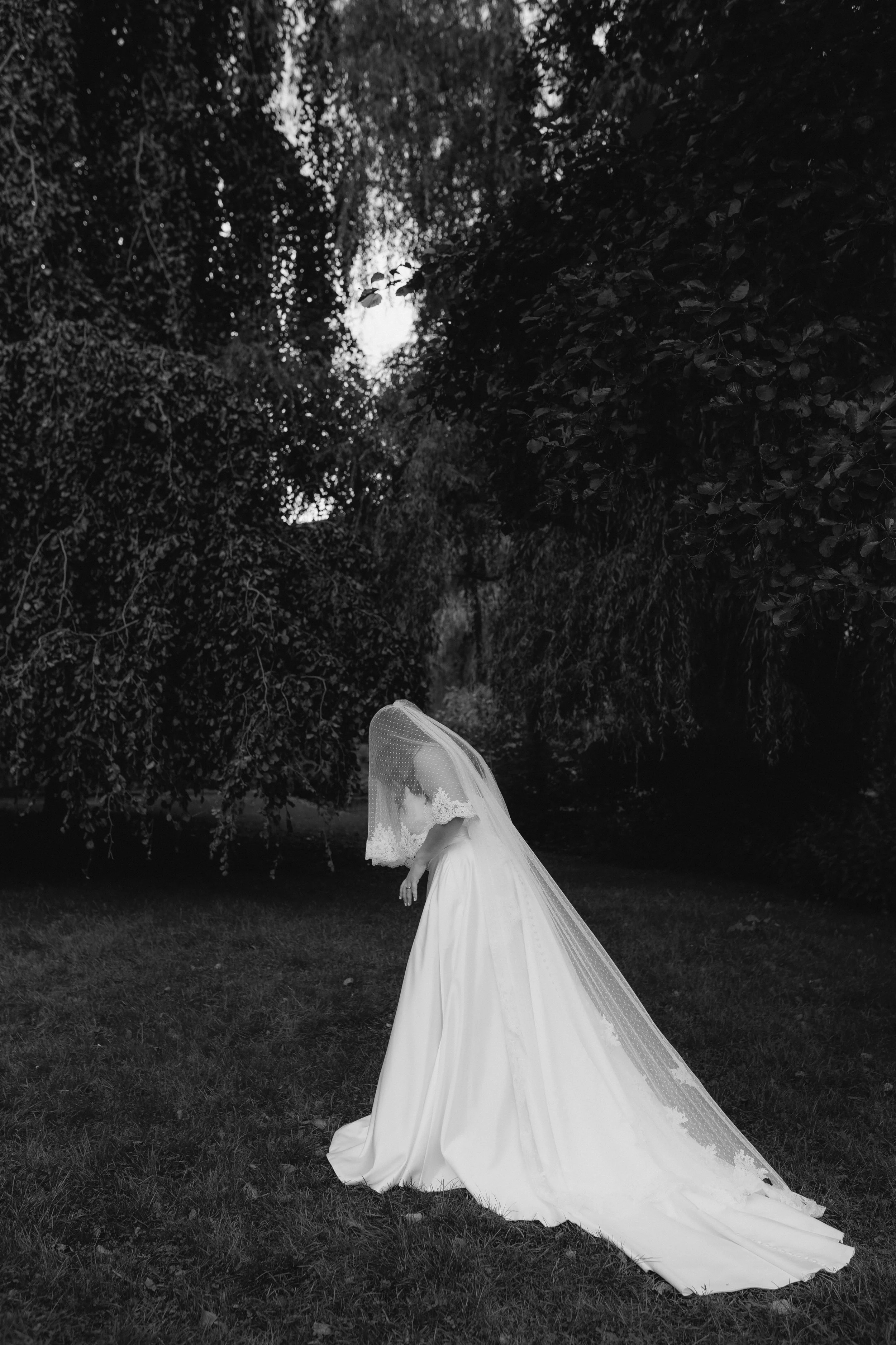 Schwarz-weiß-Foto einer Frau in Brautkleid mit Schleier, die auf Rasen unter Bäumen steht, mit dem Kopf leicht geneigt nach vorne.