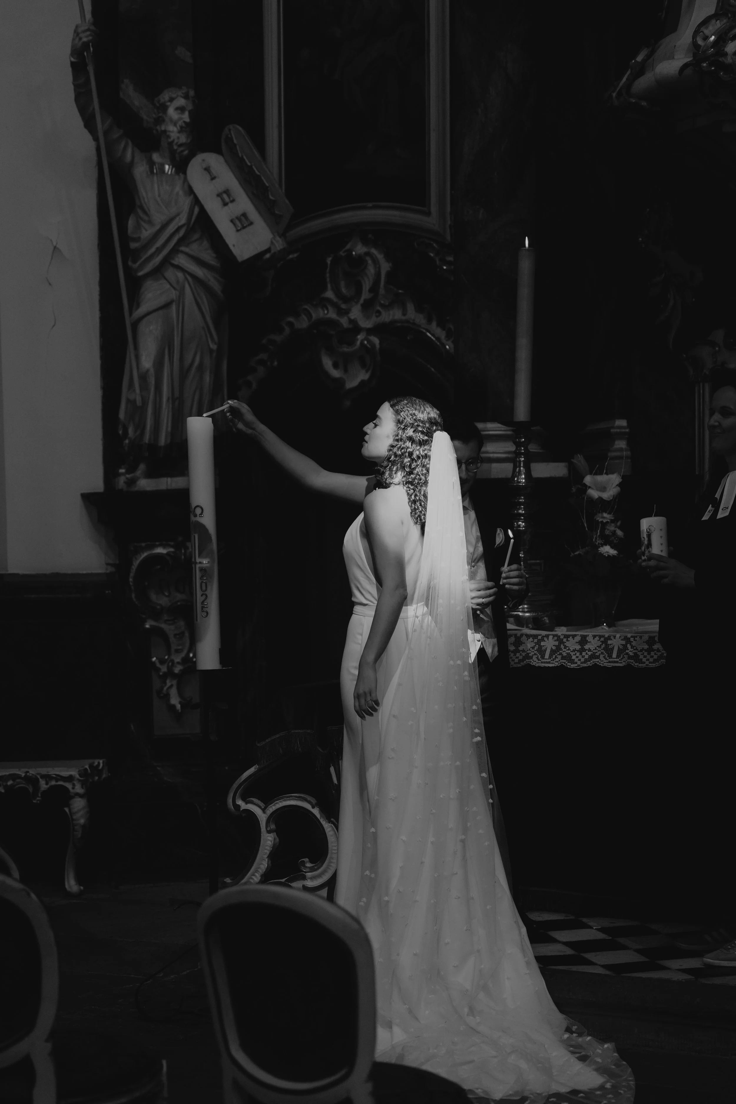 Jungfrau in Hochzeitskleid bei einer Zeremonie in einer Kirche, mit einem Priester im Hintergrund, dunkle künstlerische Inneneinrichtung, Kerzen, und einer Statue eines Heiligen.