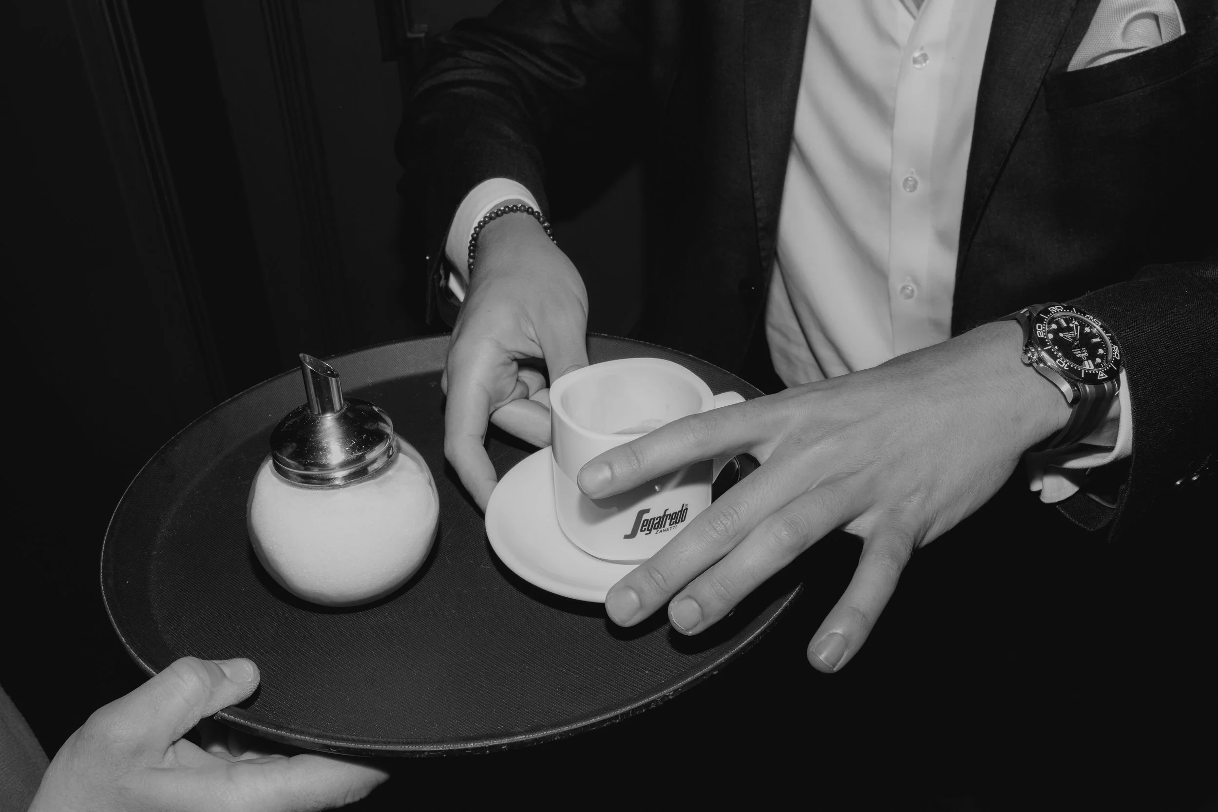 Ein Kellner serviert eine Tasse Kaffee auf einem Tablett. Der Kellner trägt eine Armbanduhr und ein Business-Outfit. Das Bild ist in Schwarz-Weiß.