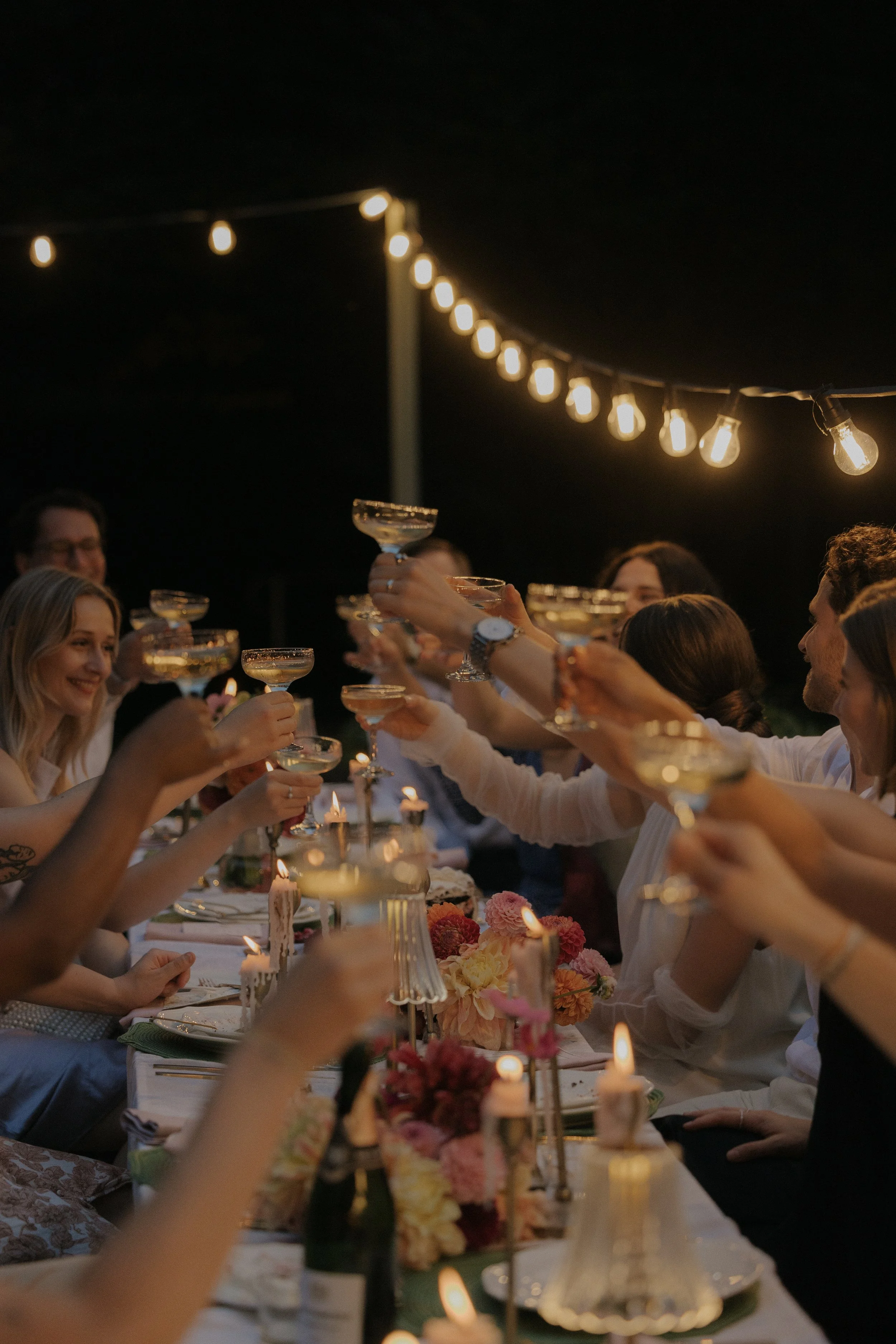 Menschen beim Anstoßen mit Gläsern während einer festlichen Abendessenparty im Freien, beleuchtet von Lichterketten.