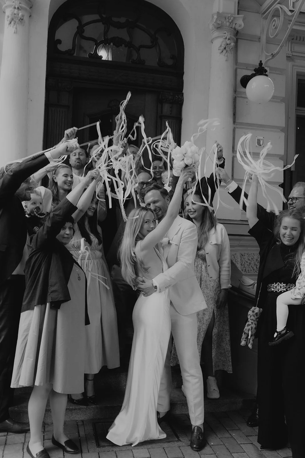 Ein Paar heiratet vor einem mehrstöckigen Gebäude, umgeben von Freunden und Familie, die mit Papierbändern und Blumen feiern.