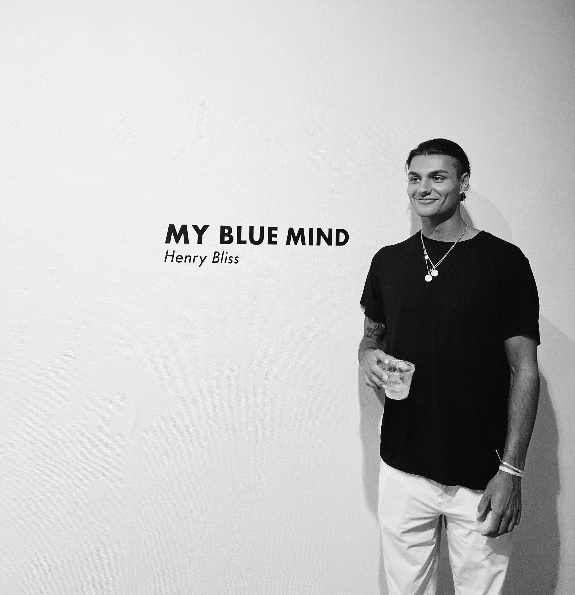 A person standing against a plain wall, holding a glass of clear liquid, wearing a black t-shirt, light-colored shorts, a necklace, and a bracelet, with tattoos on their arm. Part of the wall has text that reads, 'MY BLUE MIND' and 'Henry Bliss'.