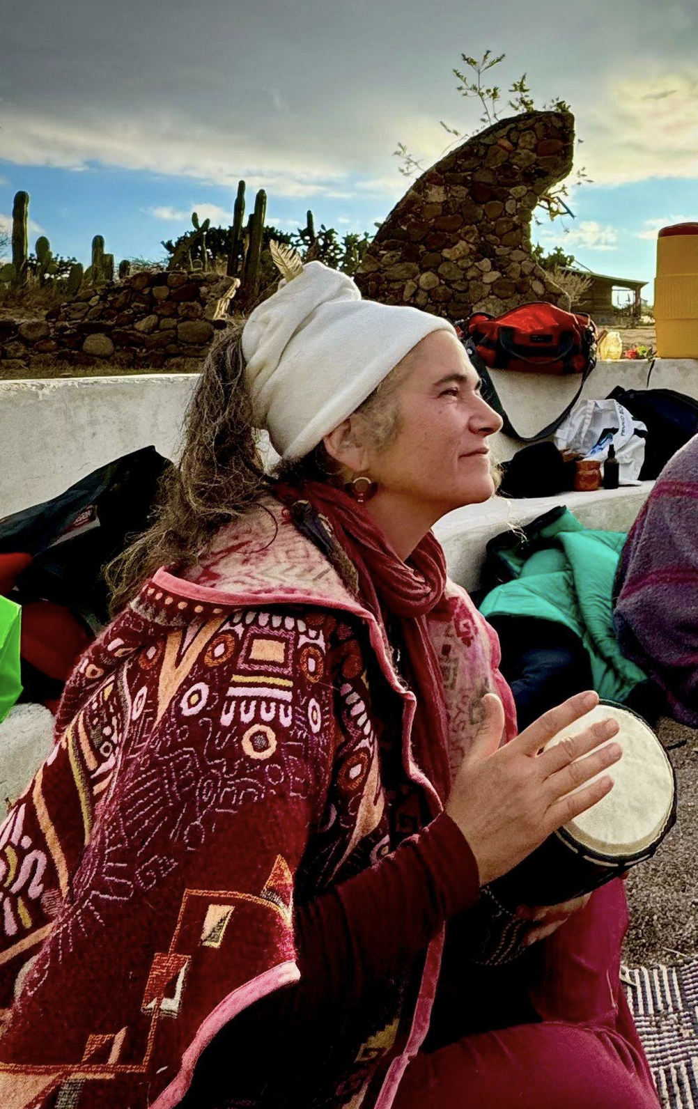 A woman wrapped in a white headscarf, sitting outdoors, playing a small drum. She is wearing a colorful patterned jacket and a red scarf. In the background, there are desert plants and a rock formation under a cloudy sky. She is happy and free.