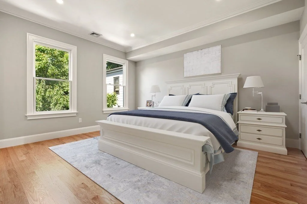 Clean and bright bedroom with white furniture, a bed with white and navy bedding, two windows showing greenery outside, and a light-colored rug on wooden floor.