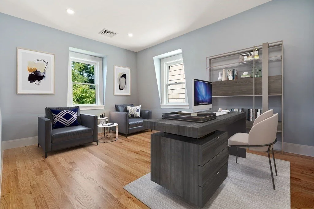 Modern home office with two gray armchairs, two windows, abstract artwork, a desk with a computer, and a bookshelf.