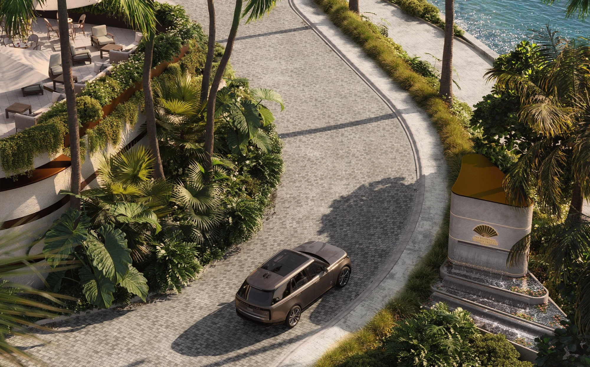Aerial view of an SUV driving on a cobblestone road lined with lush greenery, palm trees, and a waterfront. A building with a terrace is partially visible, with patio furniture and landscaping.
