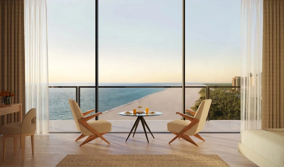 Modern living room with ocean view, featuring two lounge chairs and a small round table with drinks. Large windows provide a panoramic beach view, complemented by light curtains and a cozy rug.