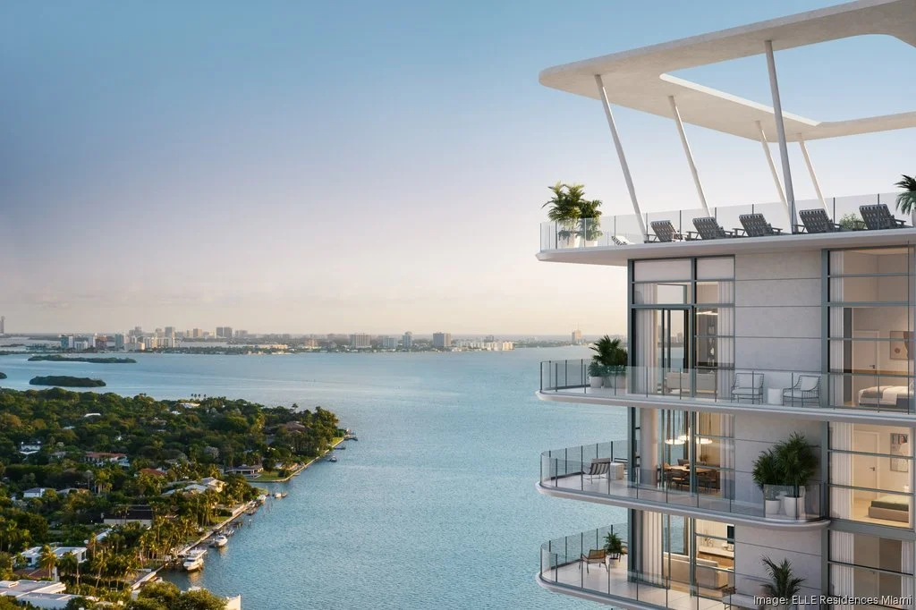 Modern luxury high-rise with balconies overlooking a body of water, featuring a city skyline and lush greenery in the background under a clear sky.