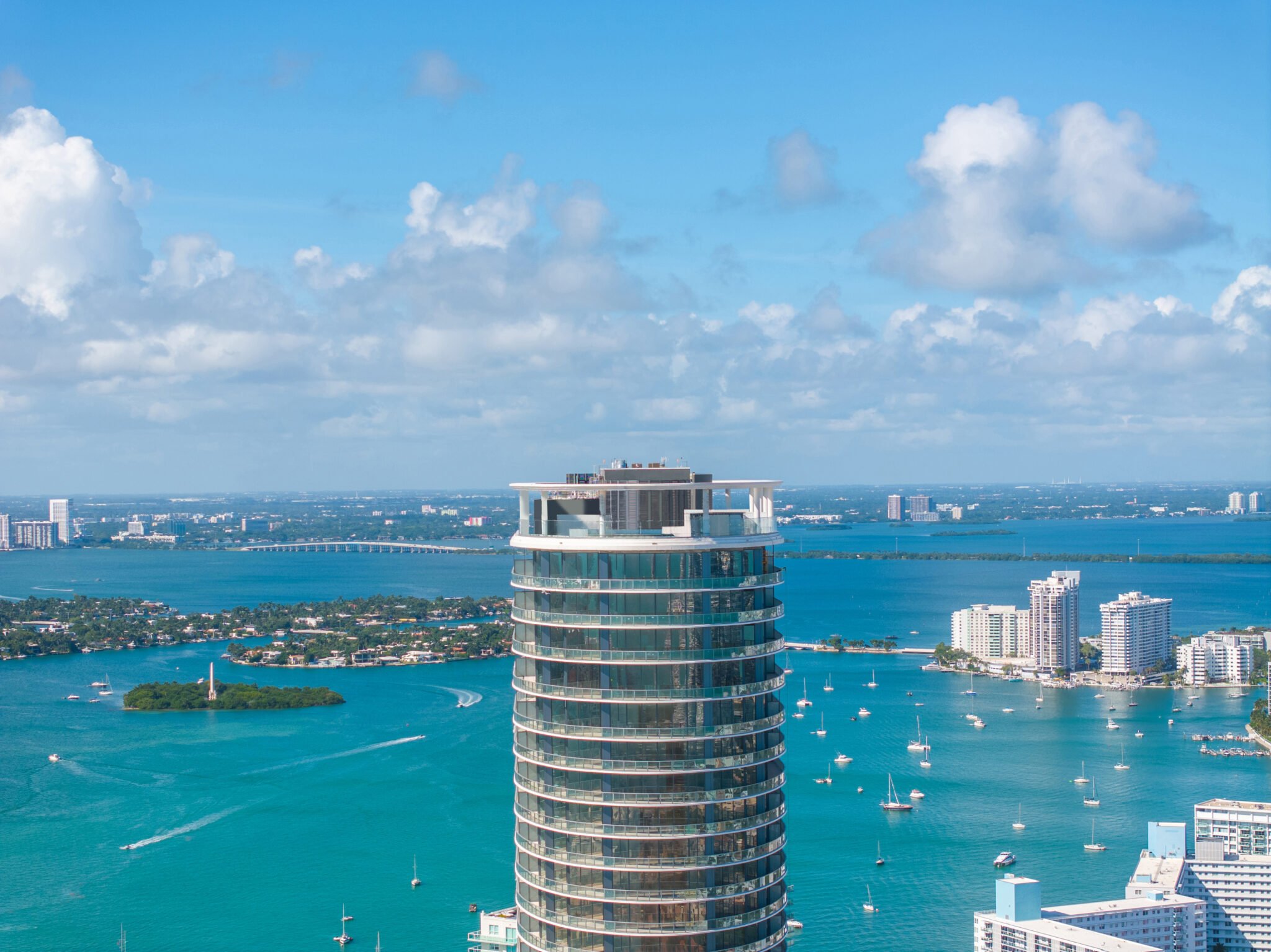 High-rise building overlooking a bay with sailboats and distant cityscape; clear blue sky with clouds.