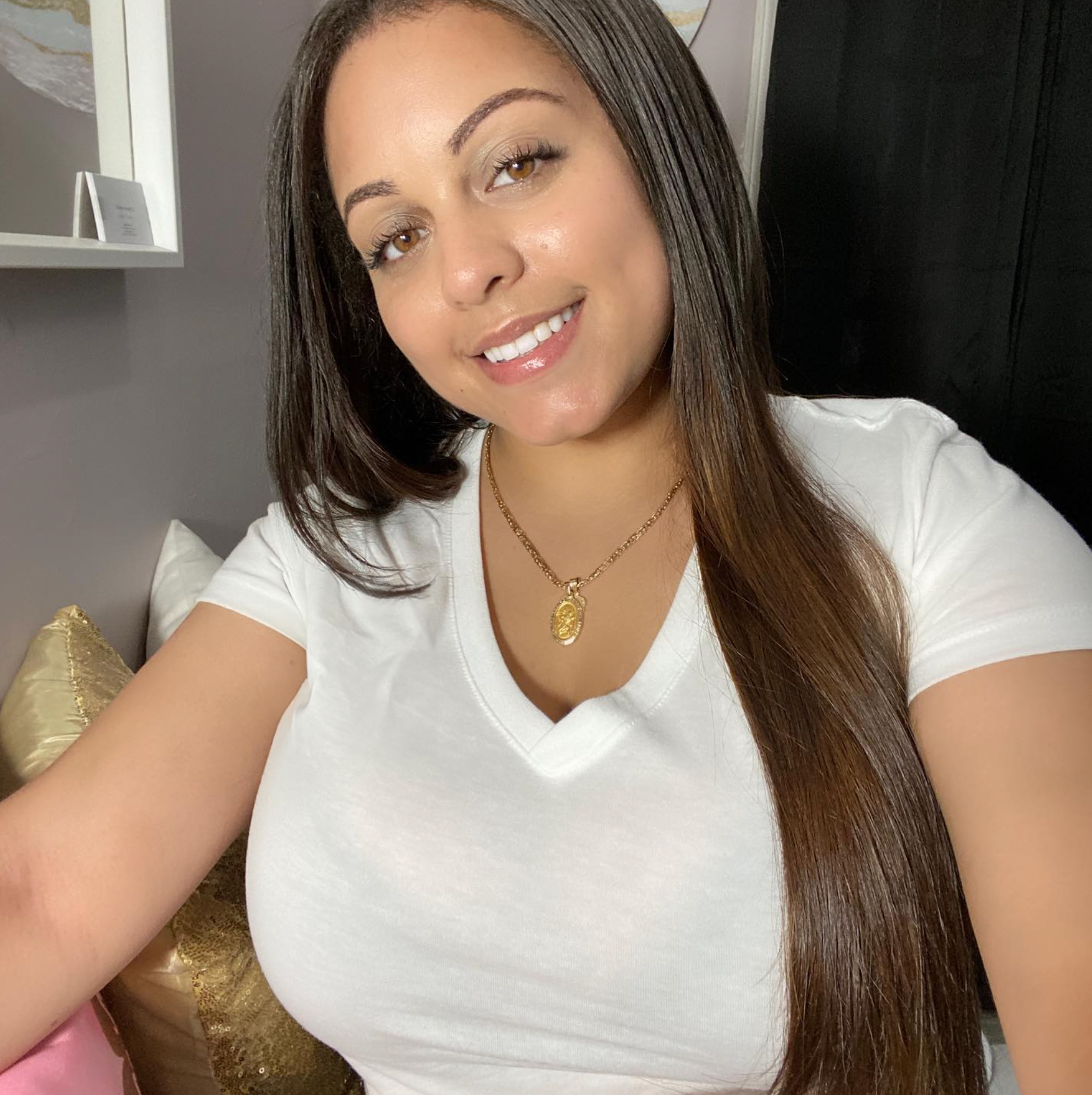 A woman with long brown hair smiling at the camera, wearing a white t-shirt and a gold necklace, sitting on a bed with gold pillows.