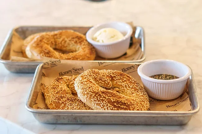 Two metal trays with sesame bagels on parchment paper, accompanied by small white cups of cream cheese and herb spread.