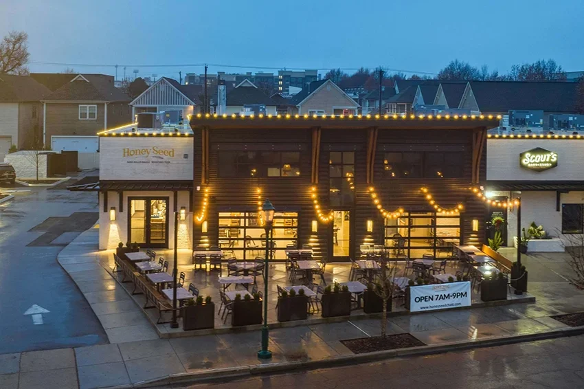 Honey Seed Chattanooga Outdoor dining patio with tables and chairs, string lights, and a lit storefront for Honey Seed bakery and Scout's bar, on a wet evening.