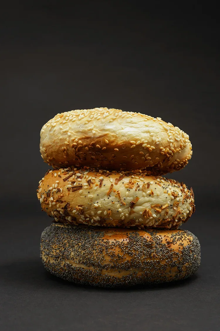 Honey Seed famous Montreal-style bagel bagels stacked vertically on a black background, each with different toppings: sesame seeds, onion, and poppy seeds.