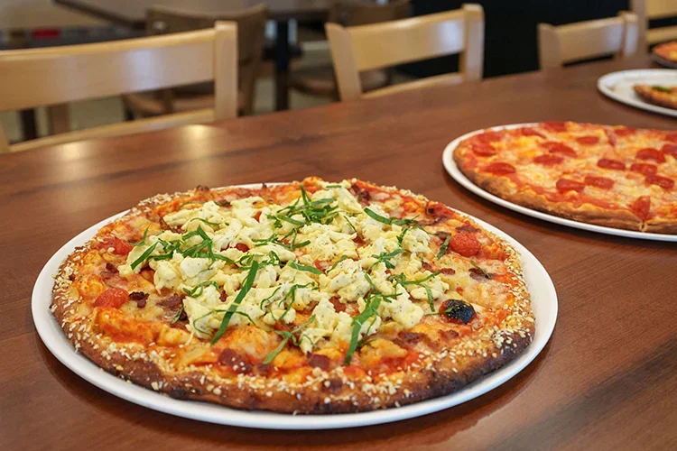 Honey Seed wood fired pizza with cheese, chicken, and herbs on a white plate, with another plain pepperoni pizza in the background, on a wooden table.
