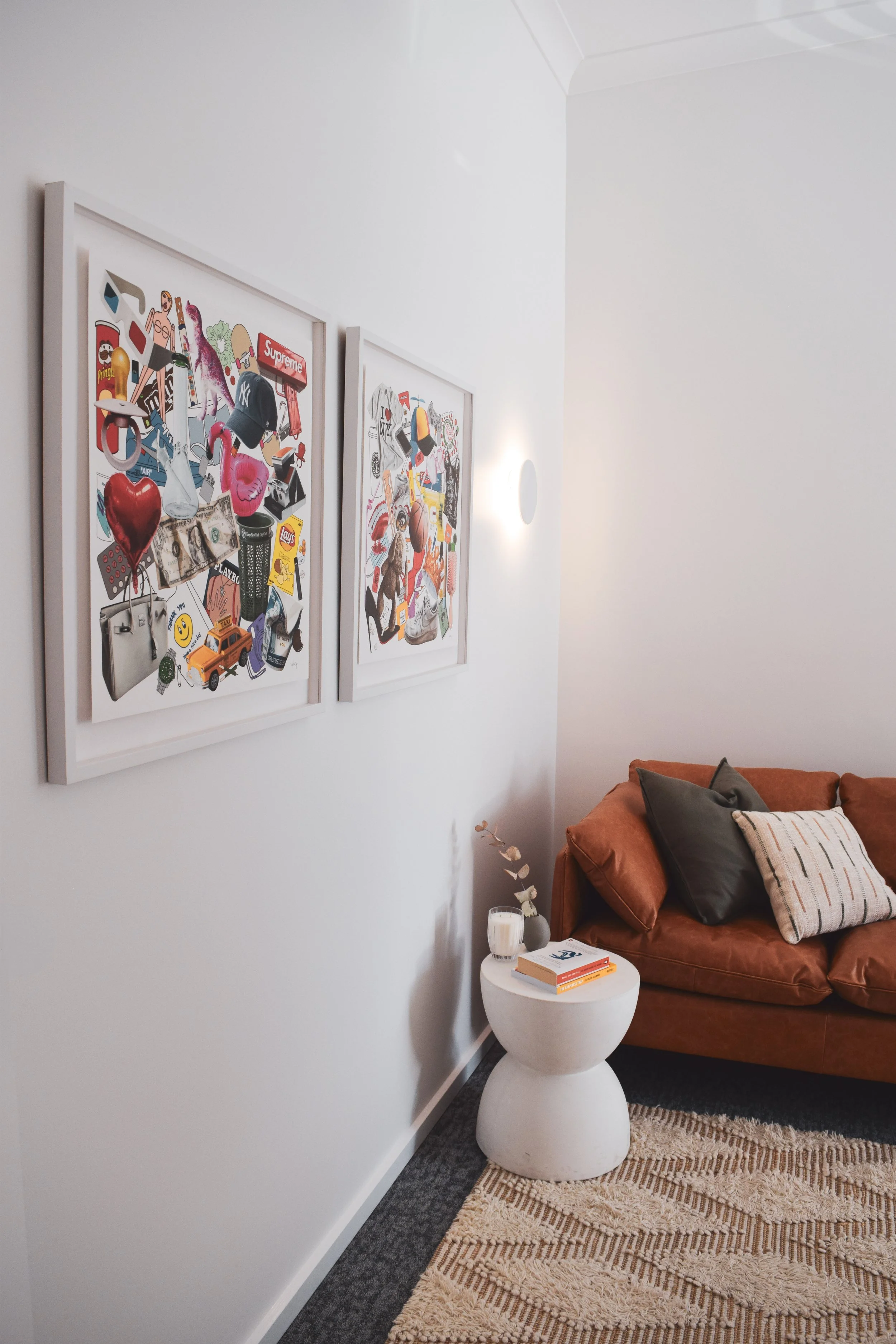 Therapy room corner with two framed collages on white wall, a brown sofa with black and beige pillows, a white round side table with books and a small vase, and a beige patterned rug.
