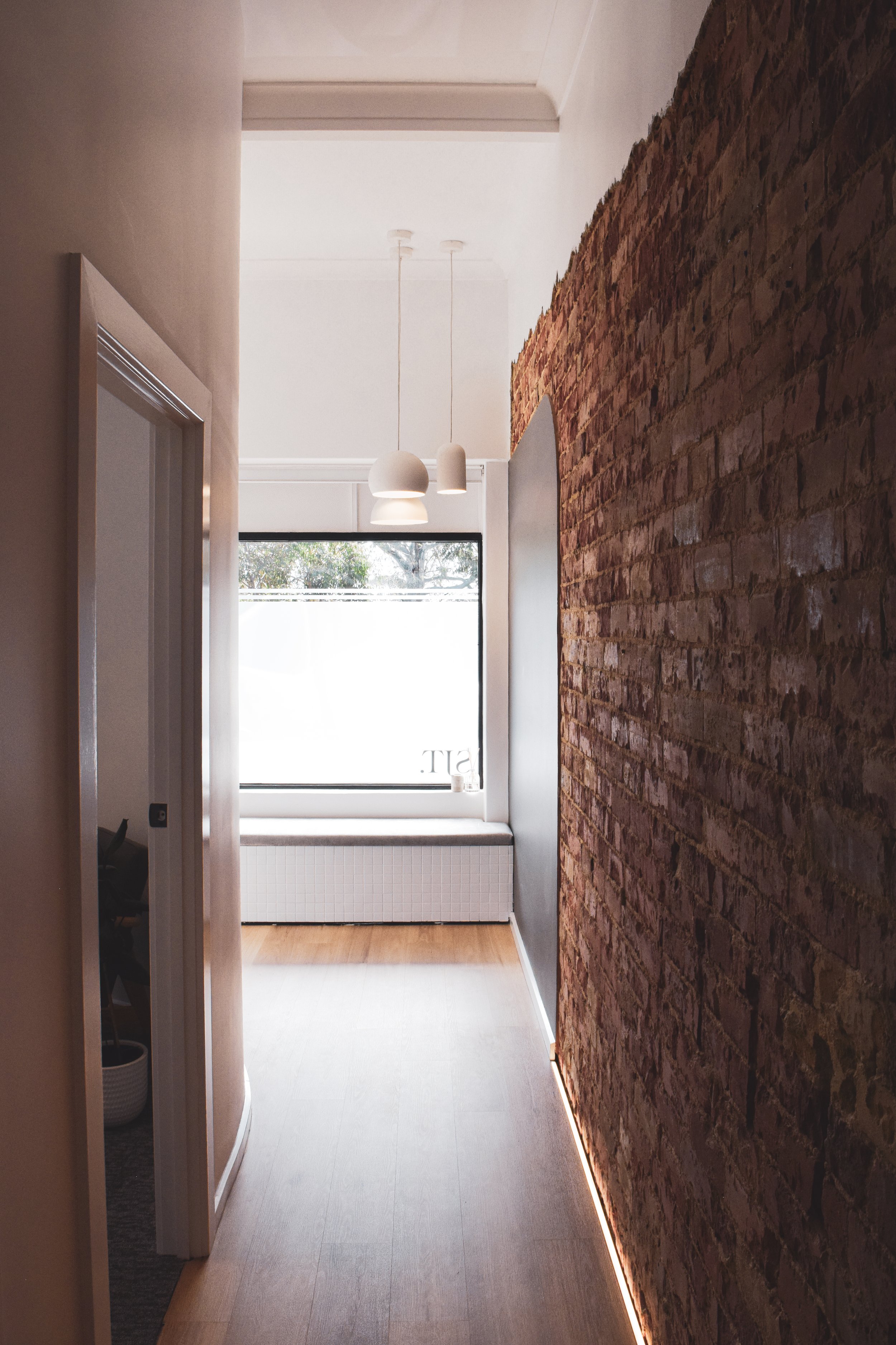 A narrow hallway with a brick wall on the right, a white wall on the left, and a large window at the end. The window has a white radiator beneath it and two pendant lights hanging from the ceiling.