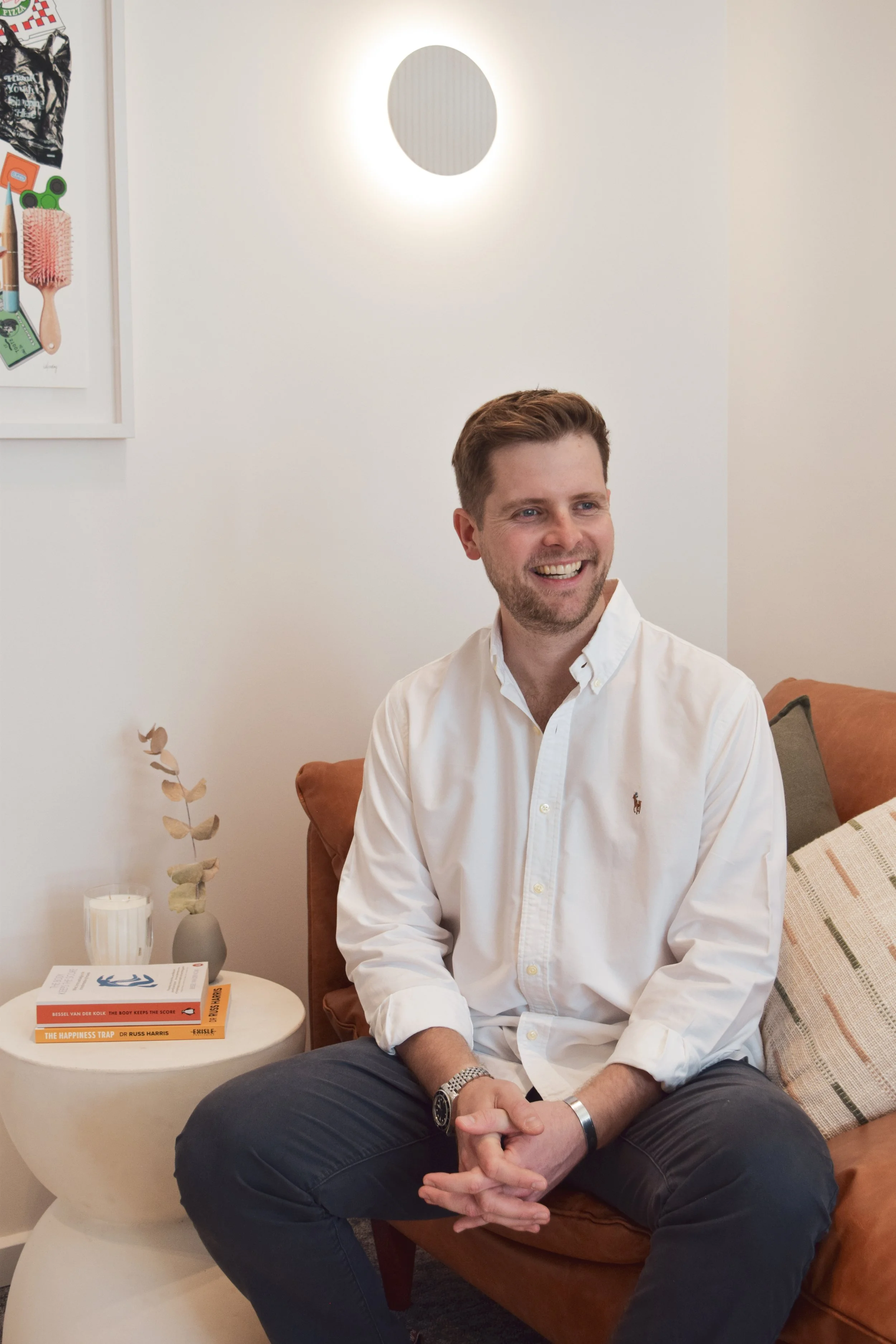 A smiling man with short blond hair and a beard, wearing a white button-up shirt and dark pants, sitting on a brown sofa in a modern, well-lit therapy room.