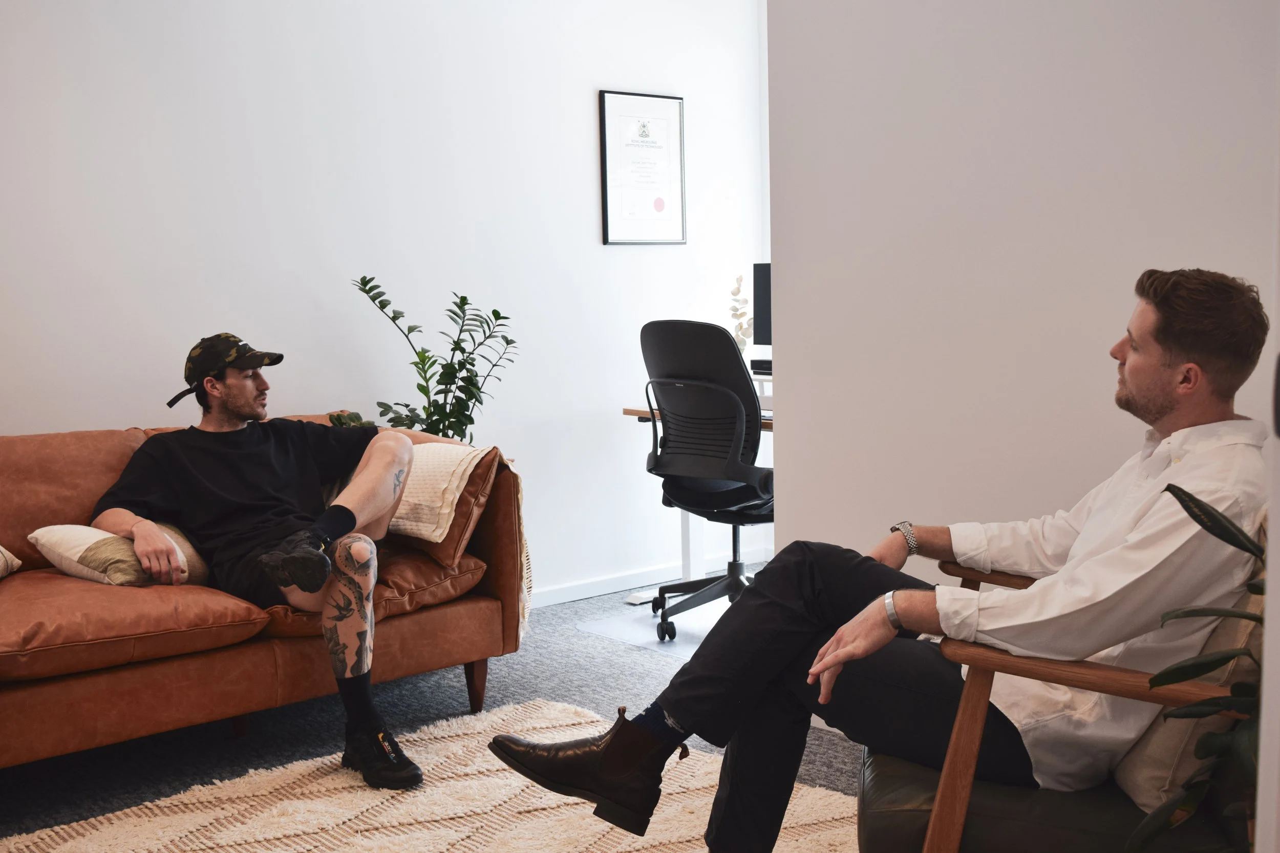 A man with tattoos sitting on a brown couch talking to a therapist sitting in a chair in a therapy session.