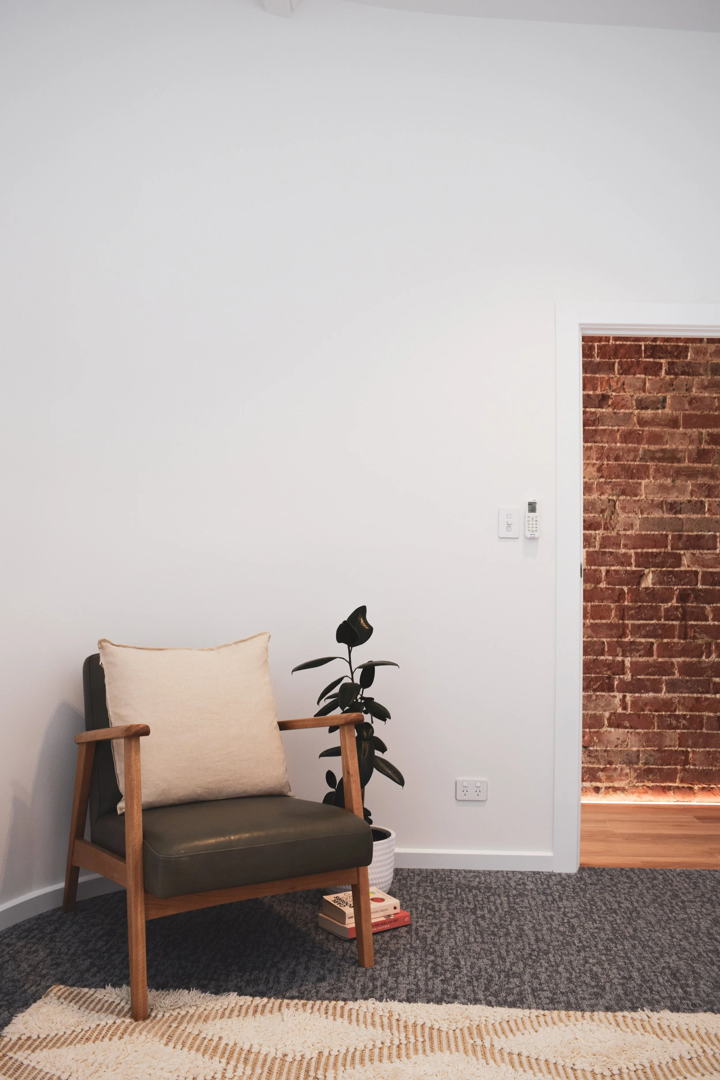 A corner of a counselling room with a wooden armchair, beige cushion, potted plant on the floor, and a small stack of books next to the plant, against a white wall and a red brick wall in the background.