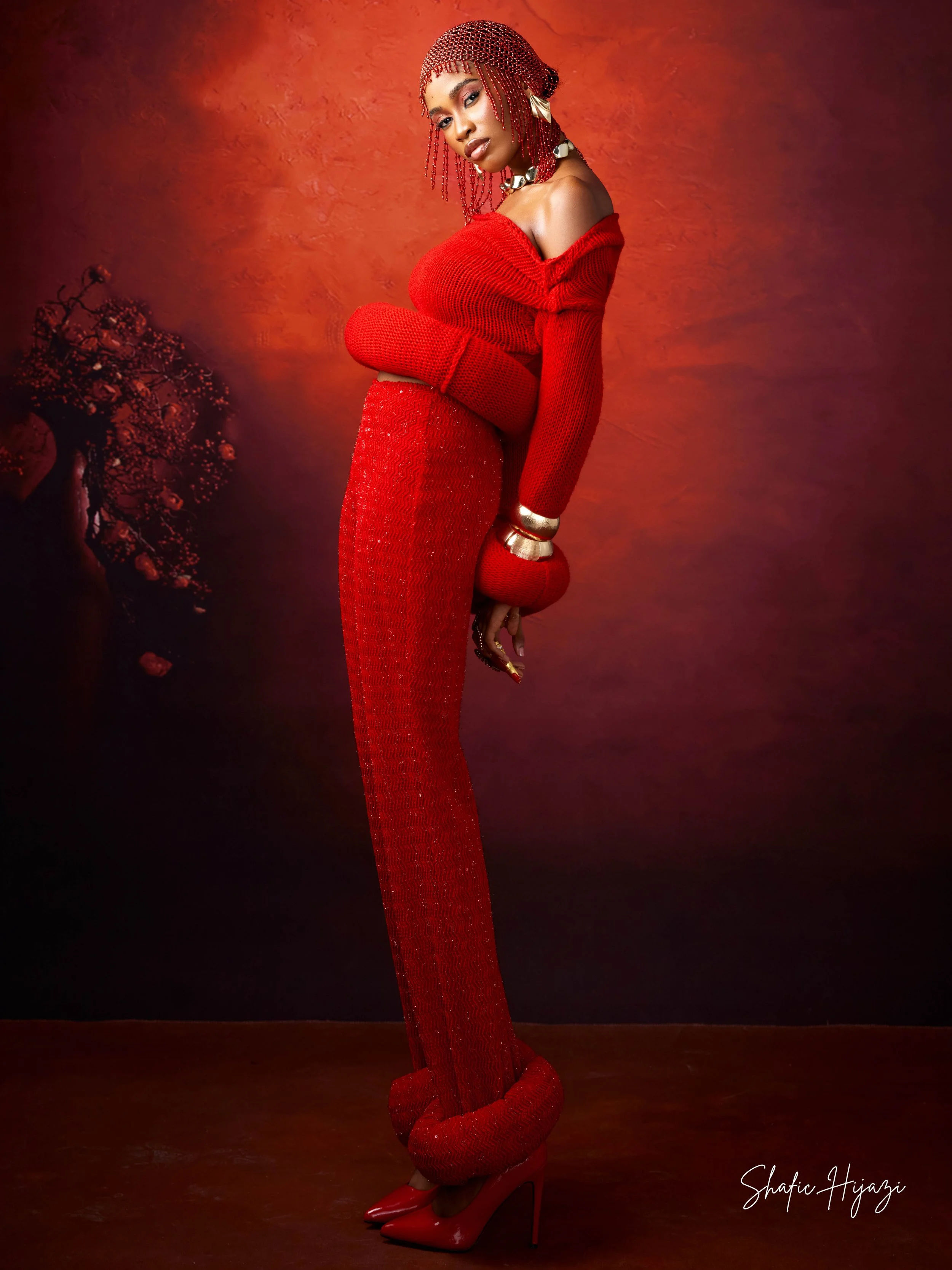 Woman wearing red high heels, a long red knit skirt, and a matching off-shoulder red top. She has braided red hair with beads, jewelry including necklaces and bangles, and is posed against a red-toned background with a painted tree on the left.