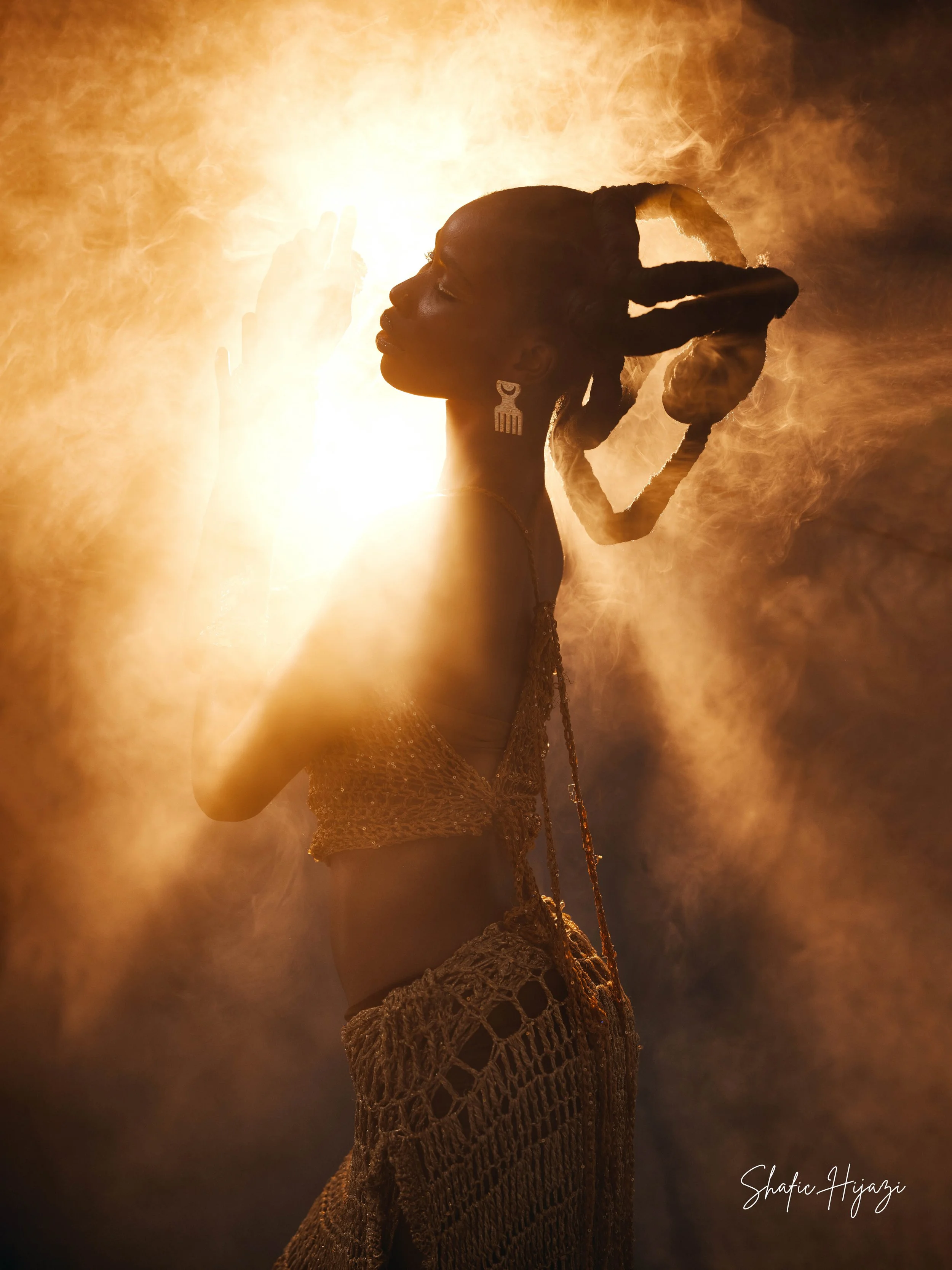 A woman with braided hair wearing earrings, a crop top, and a crochet skirt, standing in front of a bright light with smoke or fog around her.
