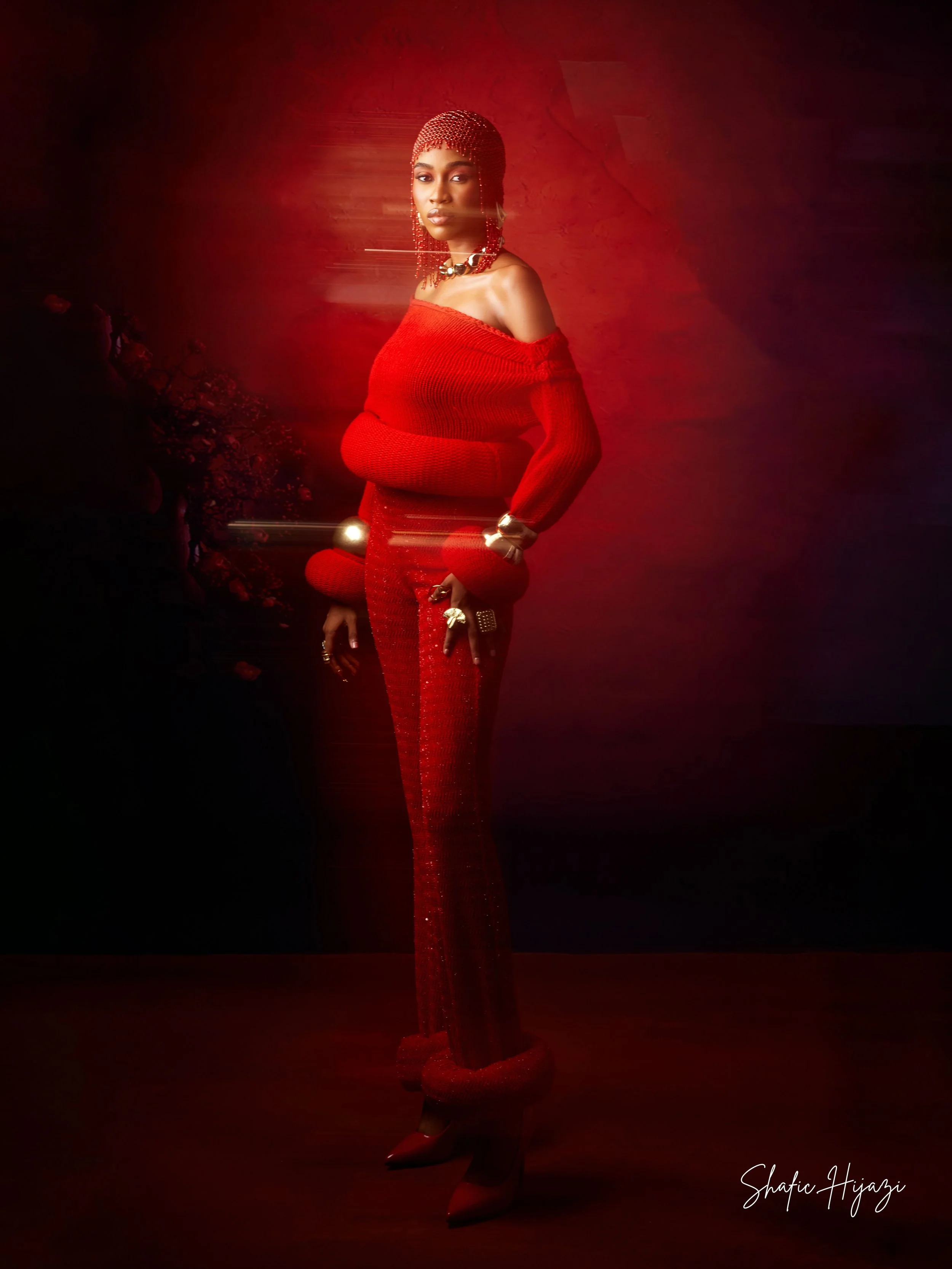 A woman in a striking, fully red outfit including a off-shoulder top and high-waisted pants, accessorized with jewelry and a beaded headscarf. The background and lighting create a dramatic effect with a red hue and motion blur.