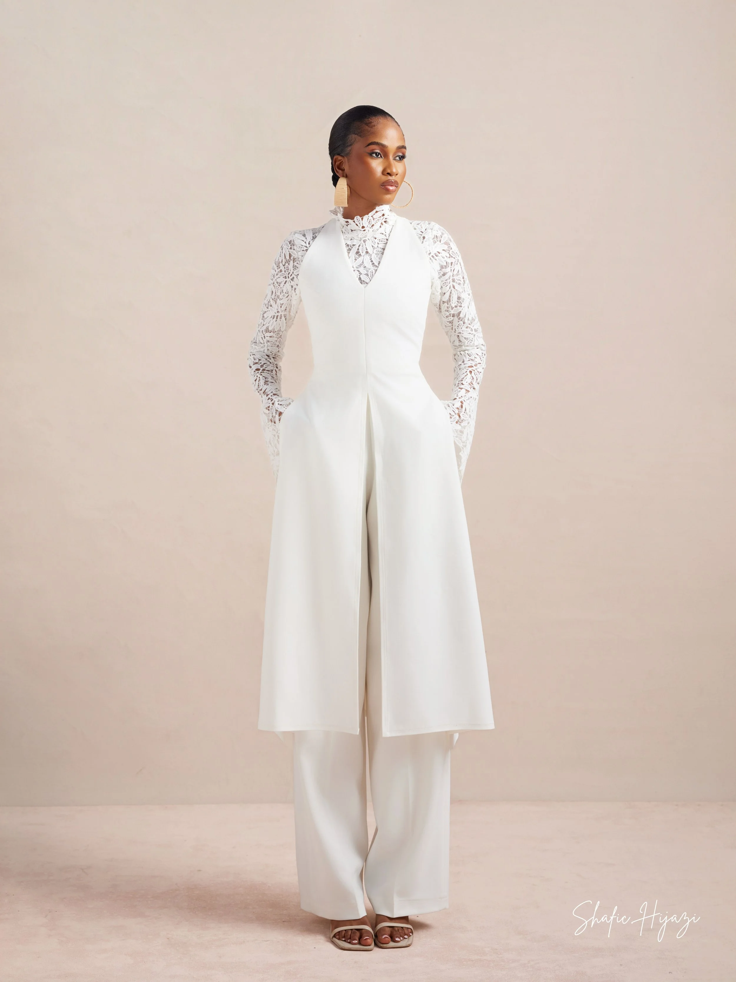 A woman wearing a white lace long-sleeve top and wide-leg pants, posing against a light beige background.