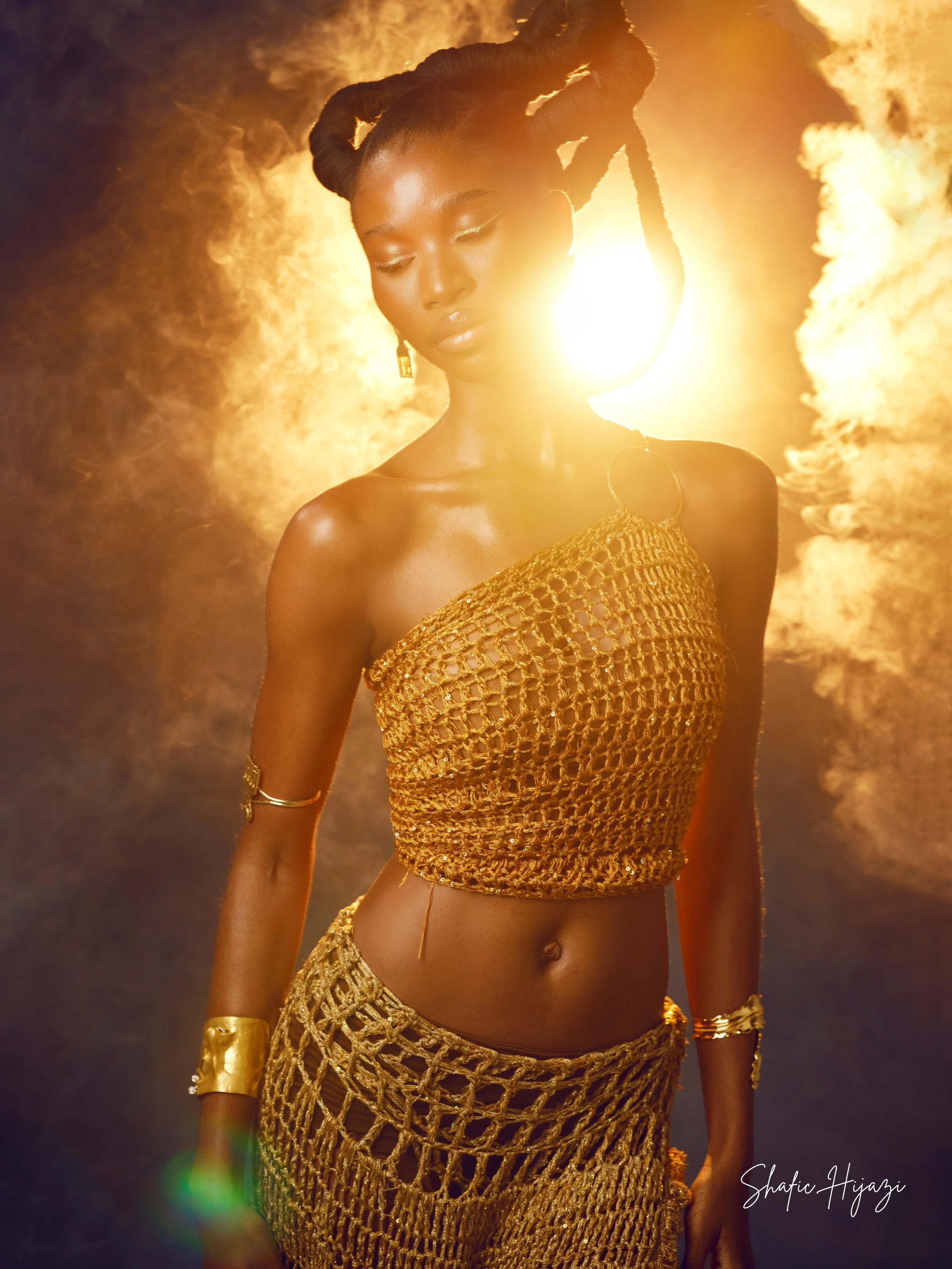 A woman dressed in gold crochet clothing with jewelry, standing in front of a fiery background with the sun shining behind her, creating a glowing effect.