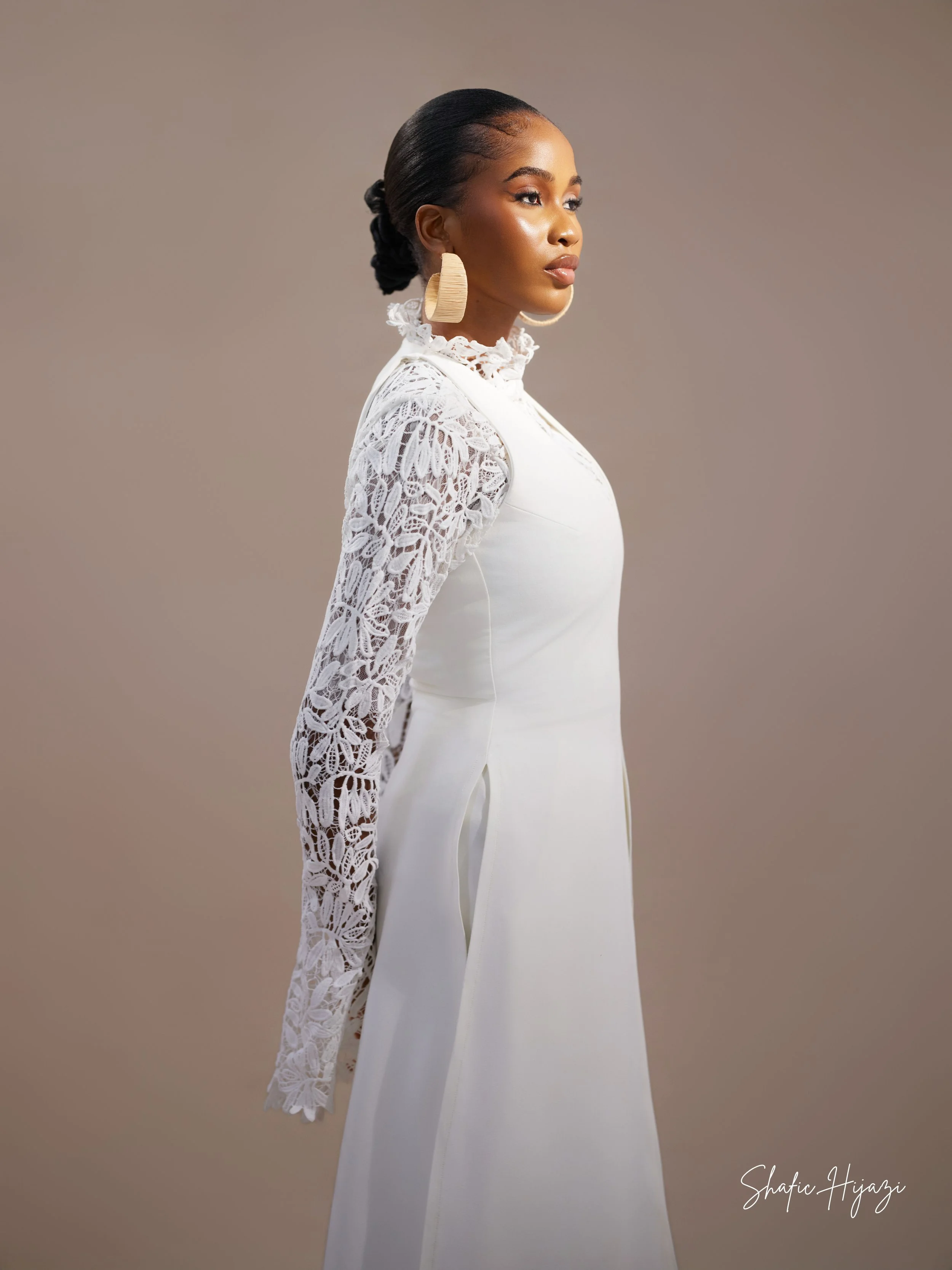 A woman in a white dress with lace sleeves and high neckline, posing against a neutral background. She has dark hair styled in a bun and is wearing large wooden earrings.