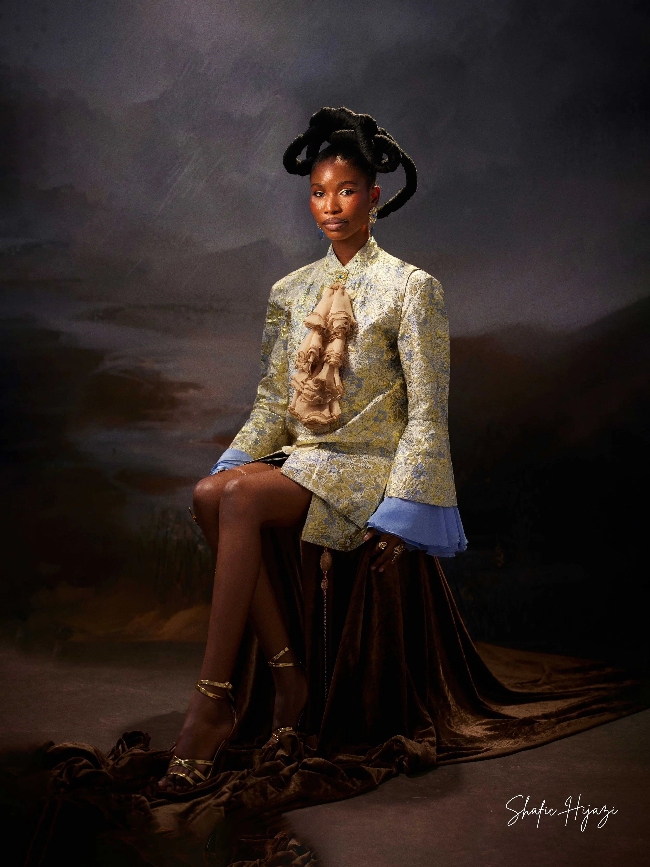 A woman in an elegant gold and light blue patterned dress with ruffled beige decoration, sitting on a dark velvet-covered stool, with her legs crossed, against a dark, dramatic background.