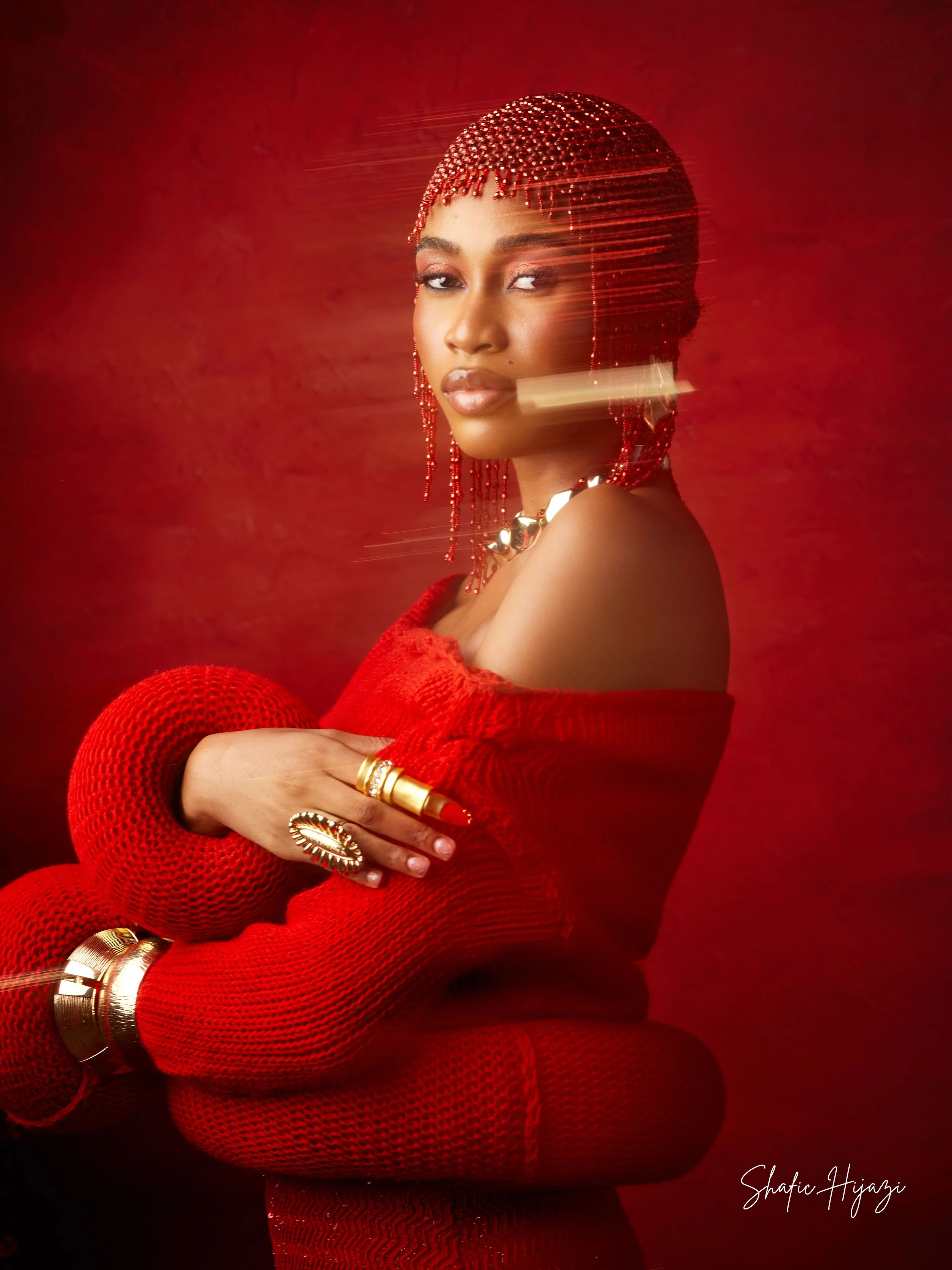 A woman dressed in a red outfit with matching red accessories, including beaded headpiece, jewelry, and long gloves, poses against a red background with a subtle texture. She has a confident expression and is looking at the camera.