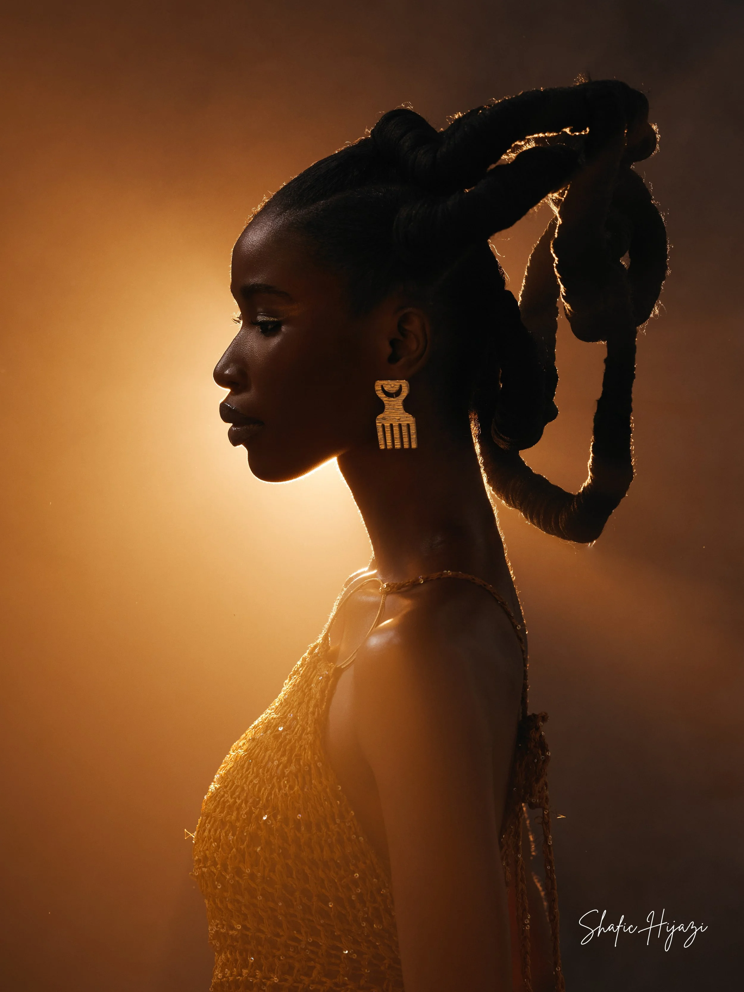Side profile of a woman with styled dreadlocks, gold jewelry, and a gold dress, illuminated by warm backlight.