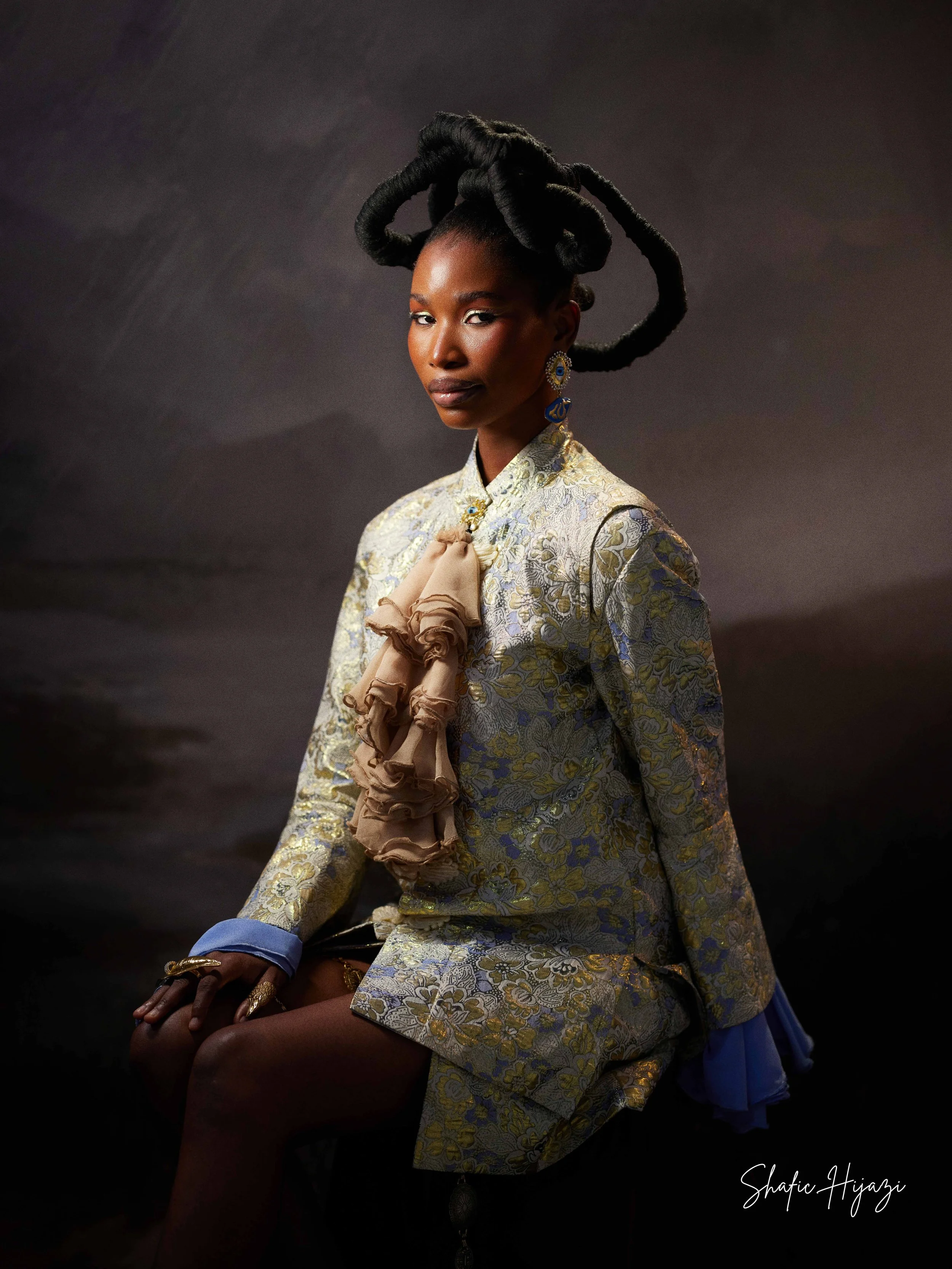 A woman with dark skin and intricate, voluminous braided hair styled in large coiled sections, sitting against a dark background. She is dressed in a richly patterned jacket with floral details, accessorized with large earrings and rings, and has a n