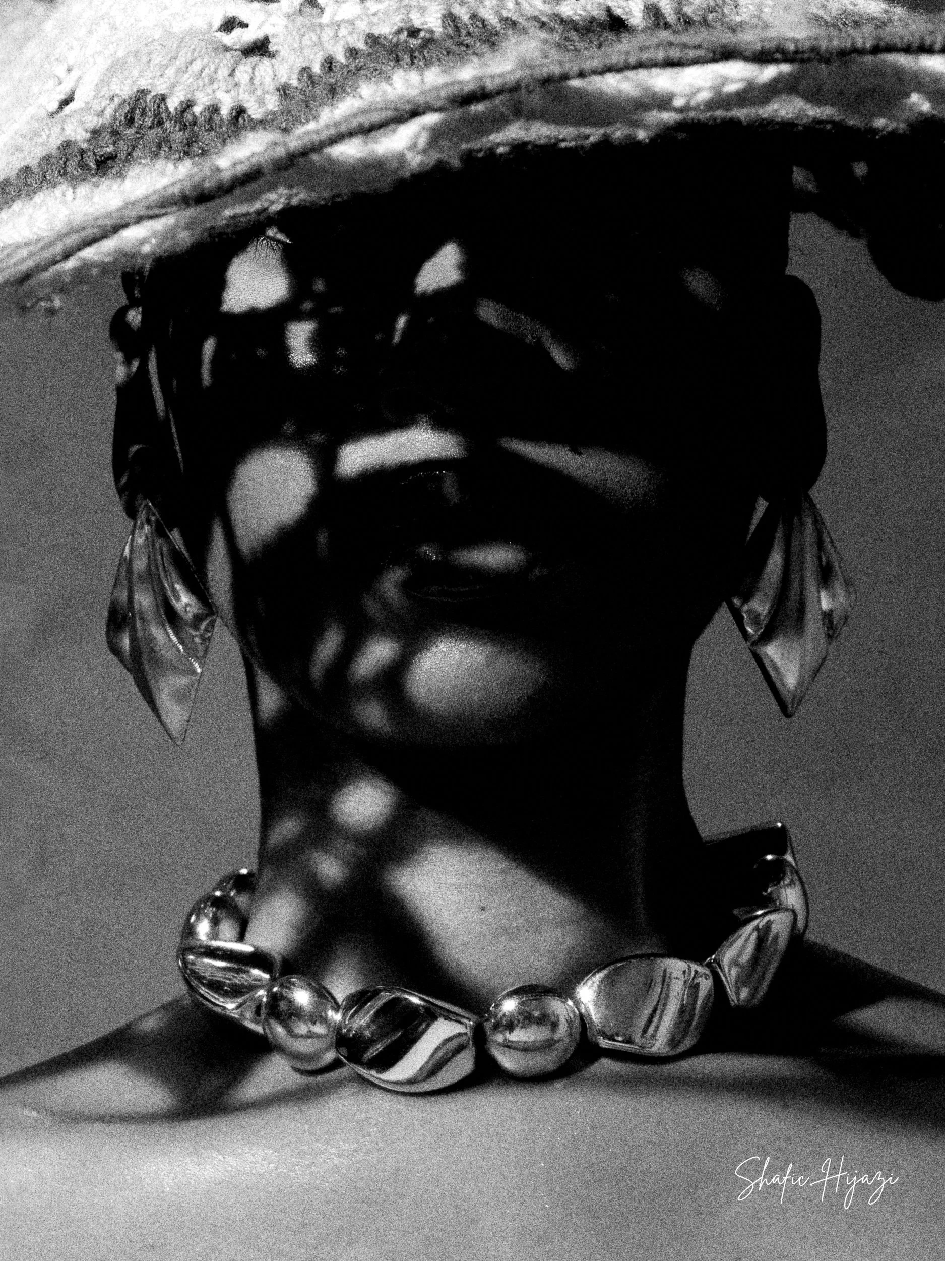 Black and white photo of a woman wearing a large hat, sunglasses, and a chunky necklace, with shadows cast over her face. The image has a signature that reads 'Shafie Hyazi.'