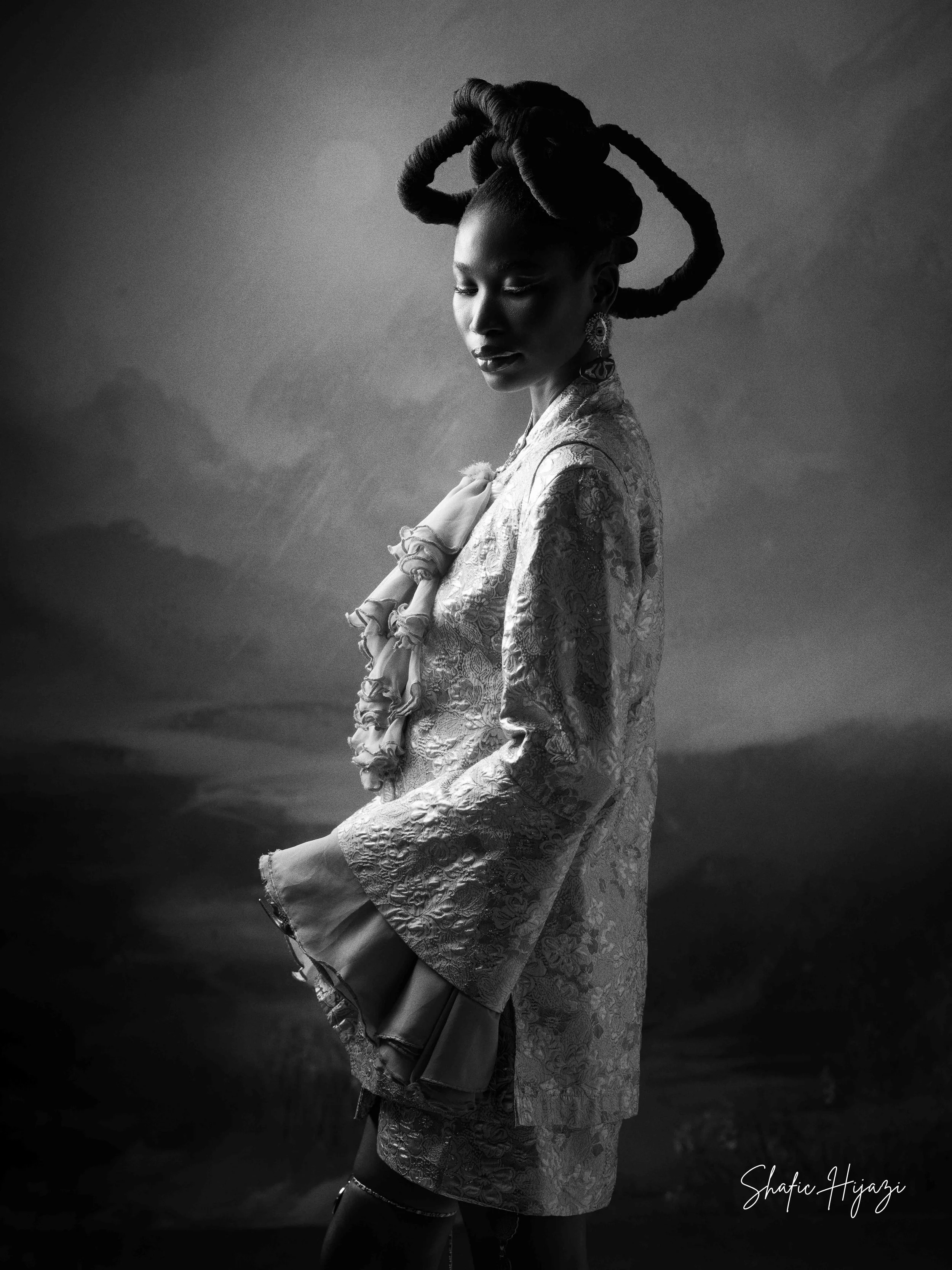 A black and white photograph of a woman with elaborate hair styled in large, sculptural twists, wearing traditional patterned clothing and jewelry, standing in front of a cloudy sky background.