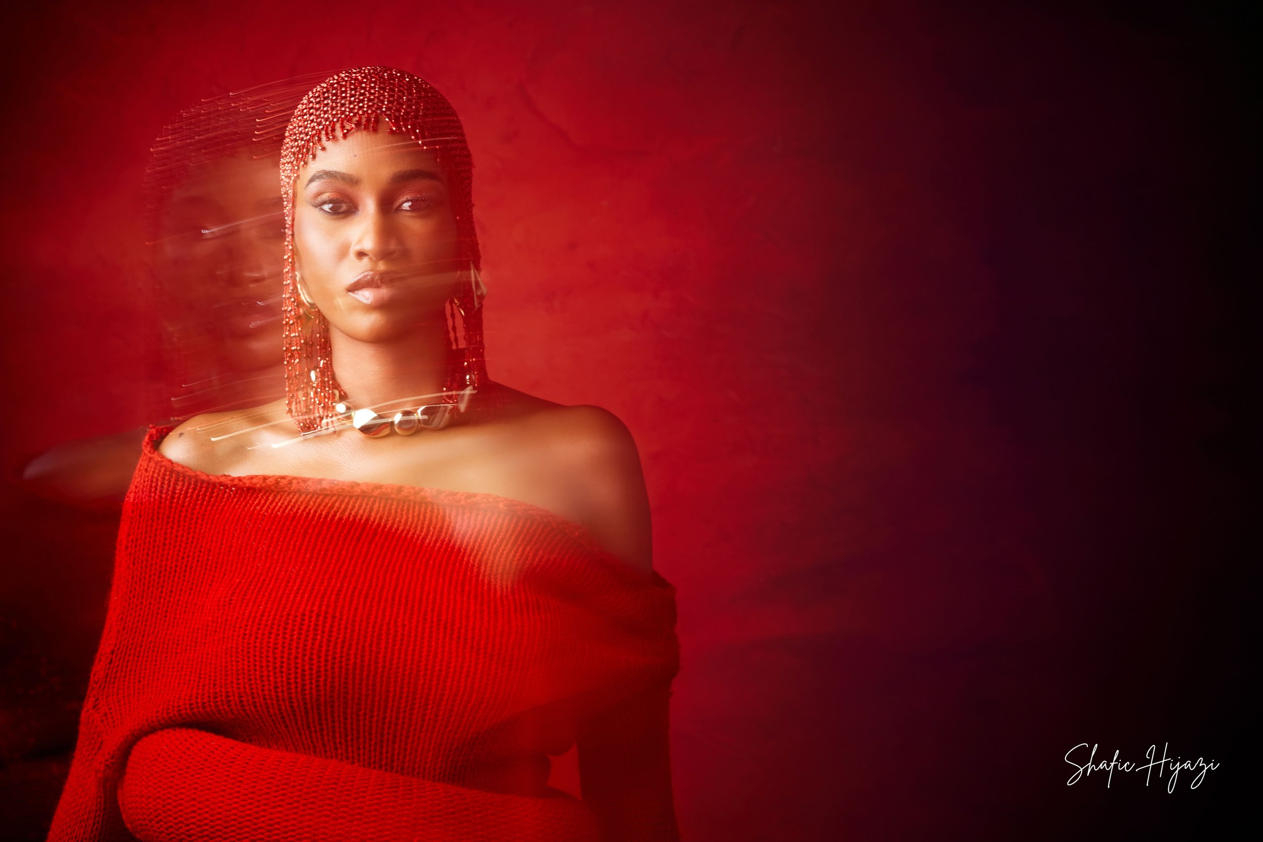 A woman with long braided hair, wearing red jewelry and a red off-shoulder sweater, standing in front of a red gradient background with motion blur effects.