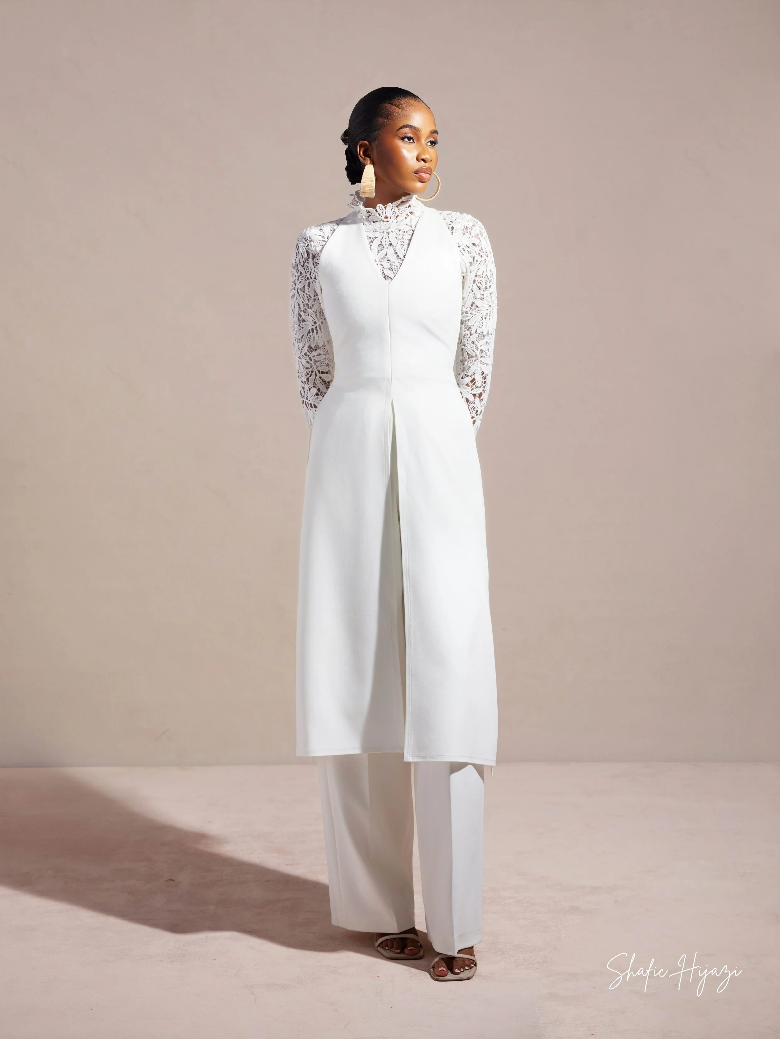 A woman wearing a white jumpsuit with lace sleeves and high heels, standing against a neutral background.