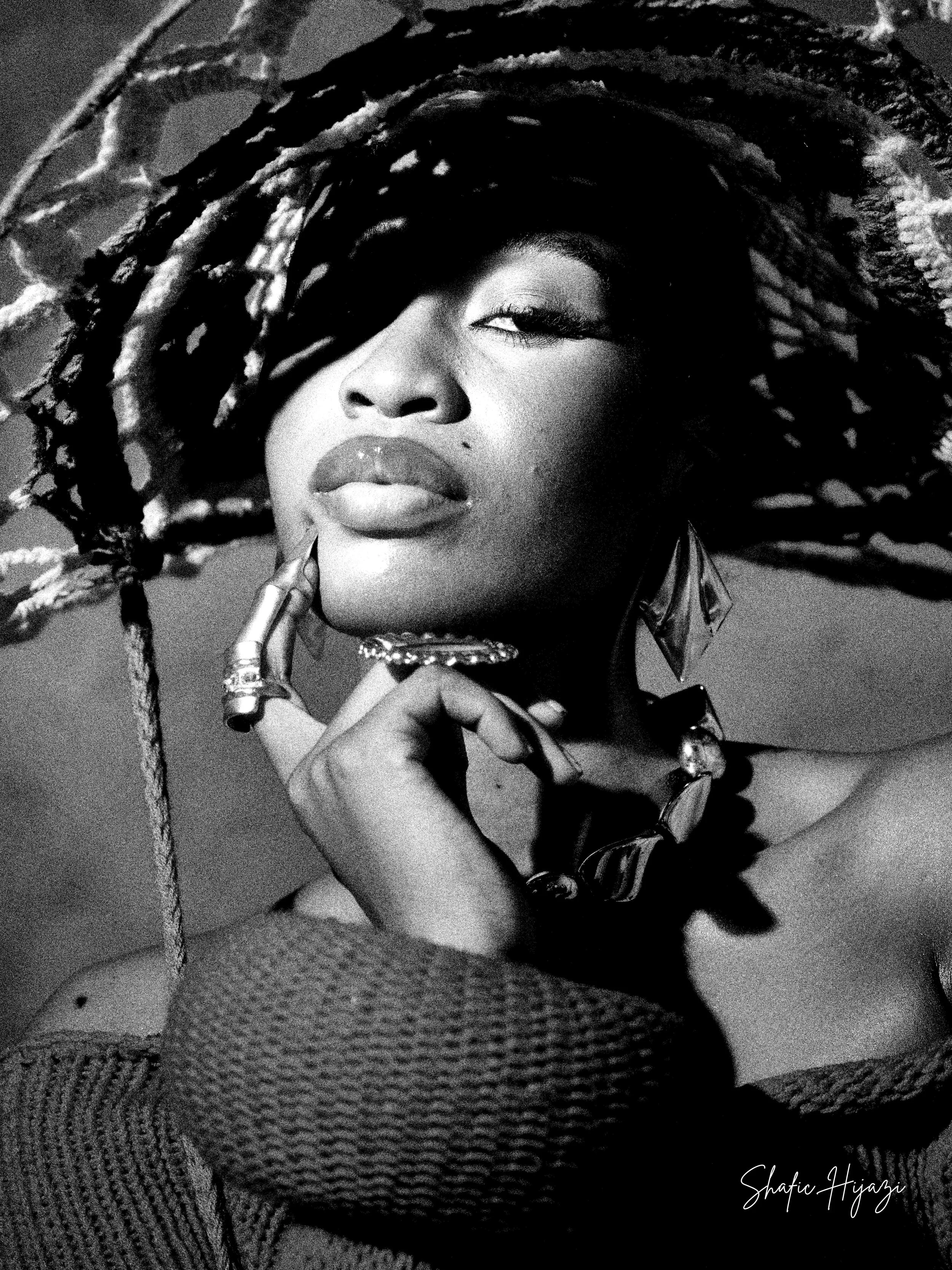 Black and white portrait of a woman with styled hair, wearing large earrings, jewelry, and a sweater, touching her face with her hand, looking at the camera with one eye closed.