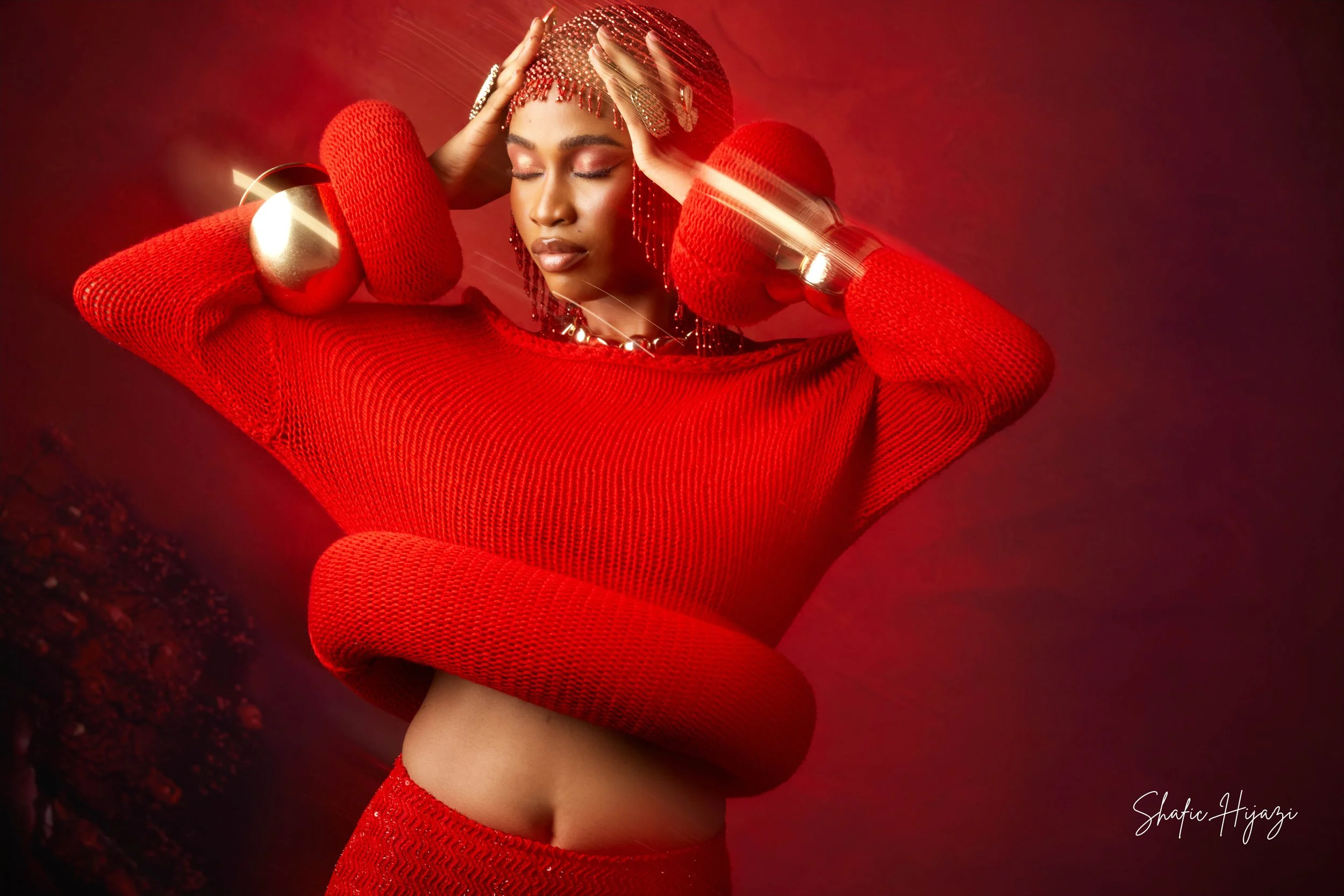 A woman with closed eyes wears a red knitted sweater with matching accessories, including large cuffs, and wraps around her waist. She has matching red headgear and jewelry, set against a red background with light streaks.