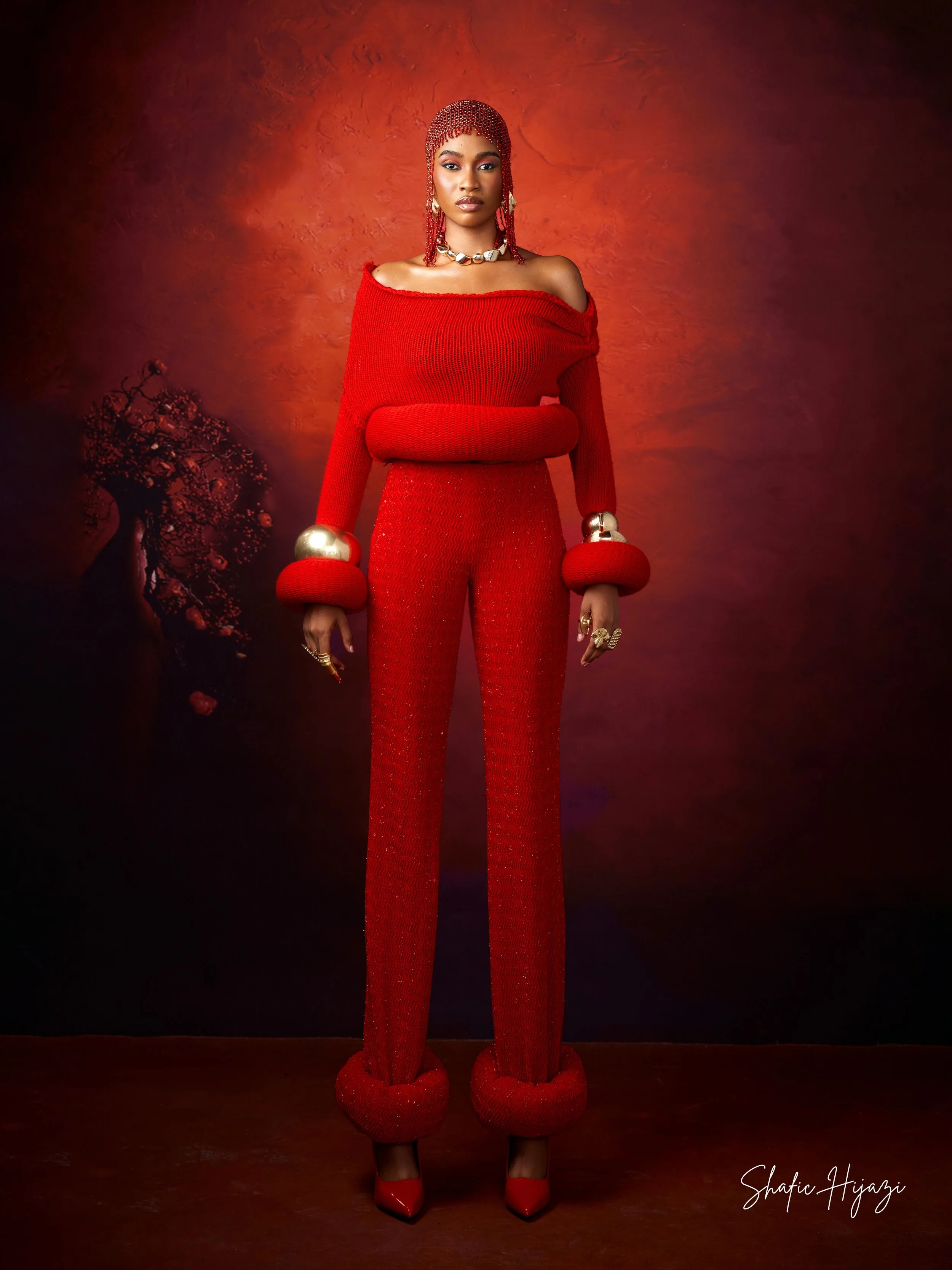 A woman dressed in a vibrant red, off-shoulder knit sweater and matching pants with pompom details at the cuffs and ankles, standing against a red abstract background. She is accessorized with jewelry including rings, a necklace, earrings, and metall