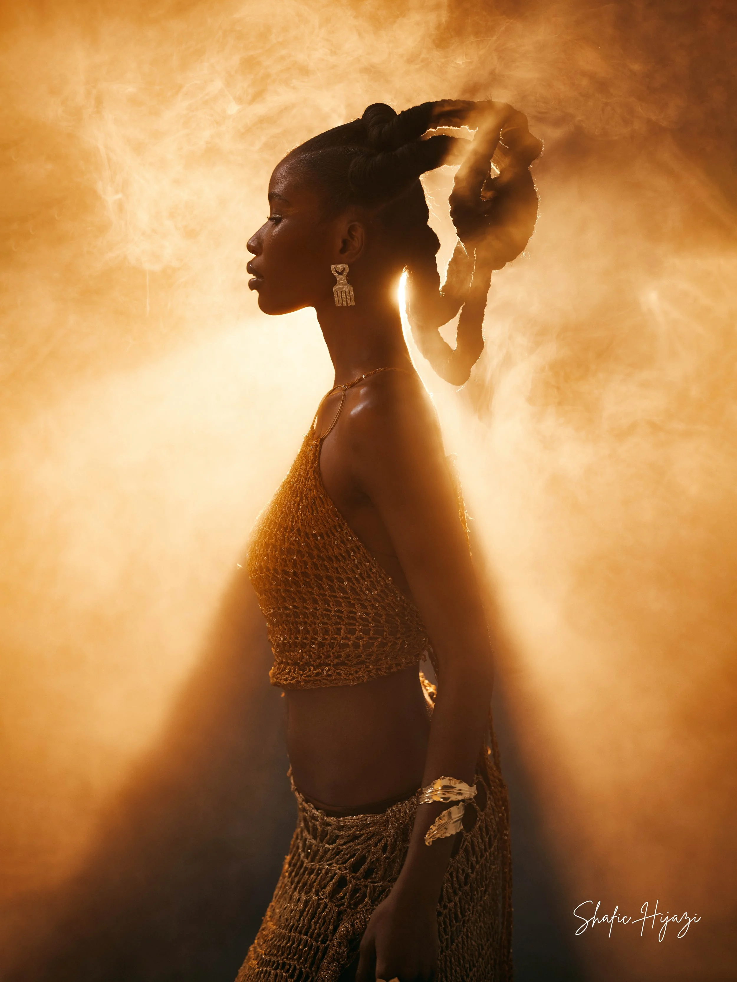 Side profile of a woman with styled hair, wearing gold jewelry and clothing, standing against a warm, glowing background.