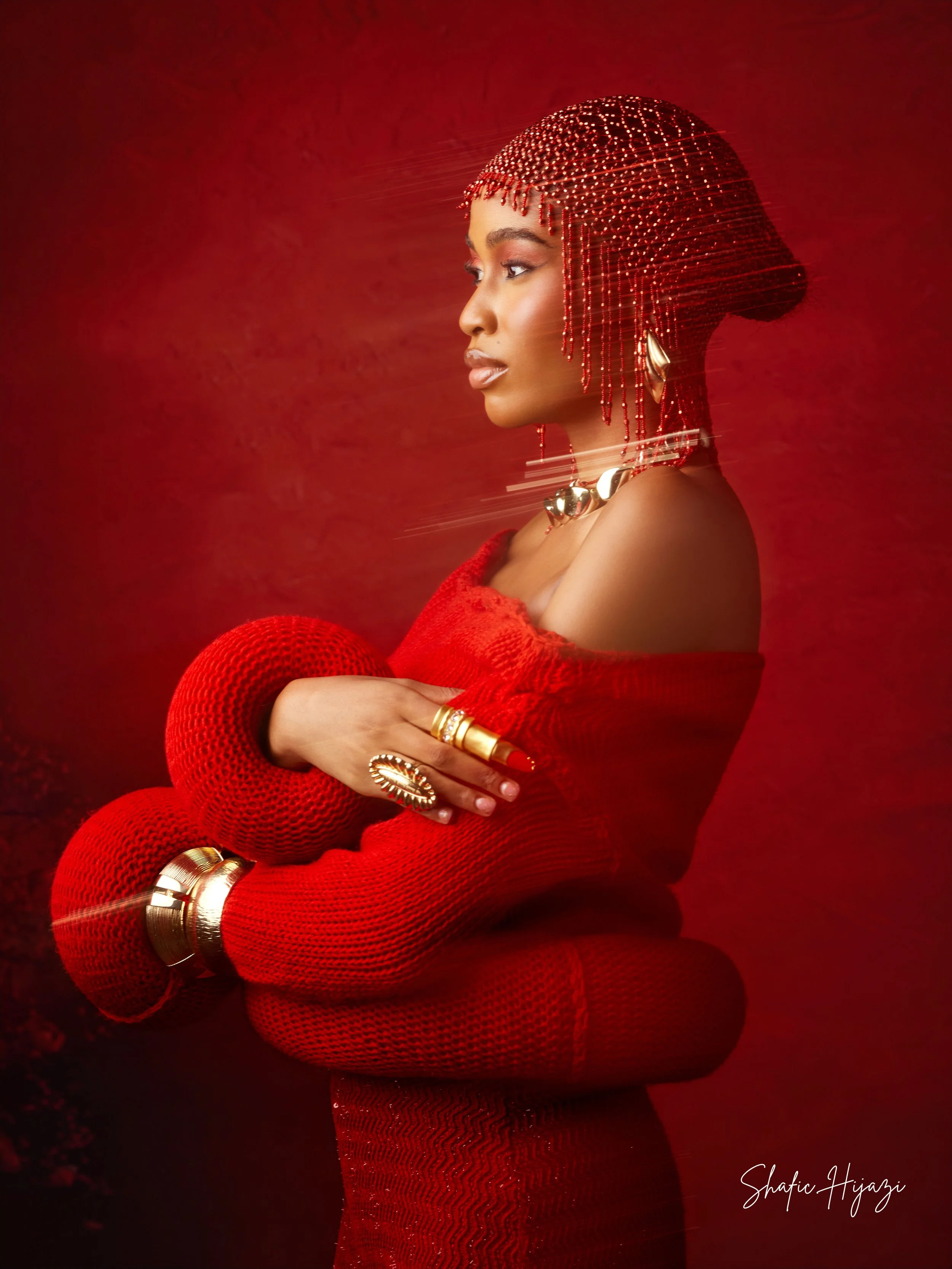 A woman in a red knit dress poses against a red background, wearing gold jewelry, including rings, bracelets, and a necklace, with a beaded red hairstyle and a matching red headpiece.