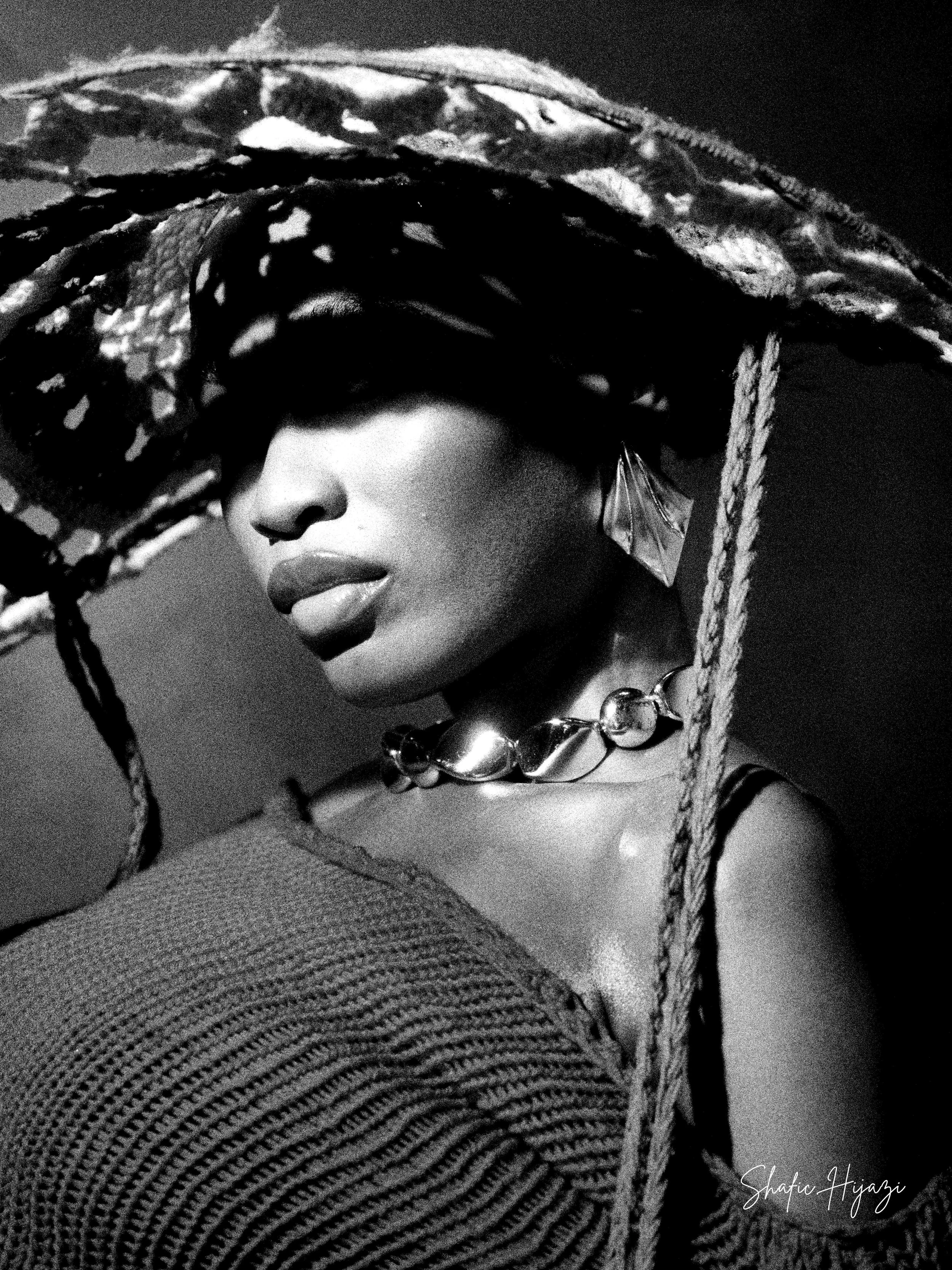 A black and white portrait of a woman wearing a large, patterned hat with a wide brim, a chunky necklace, and an earring. She has full lips and partially covered eyes, with a textured top visible on one shoulder.