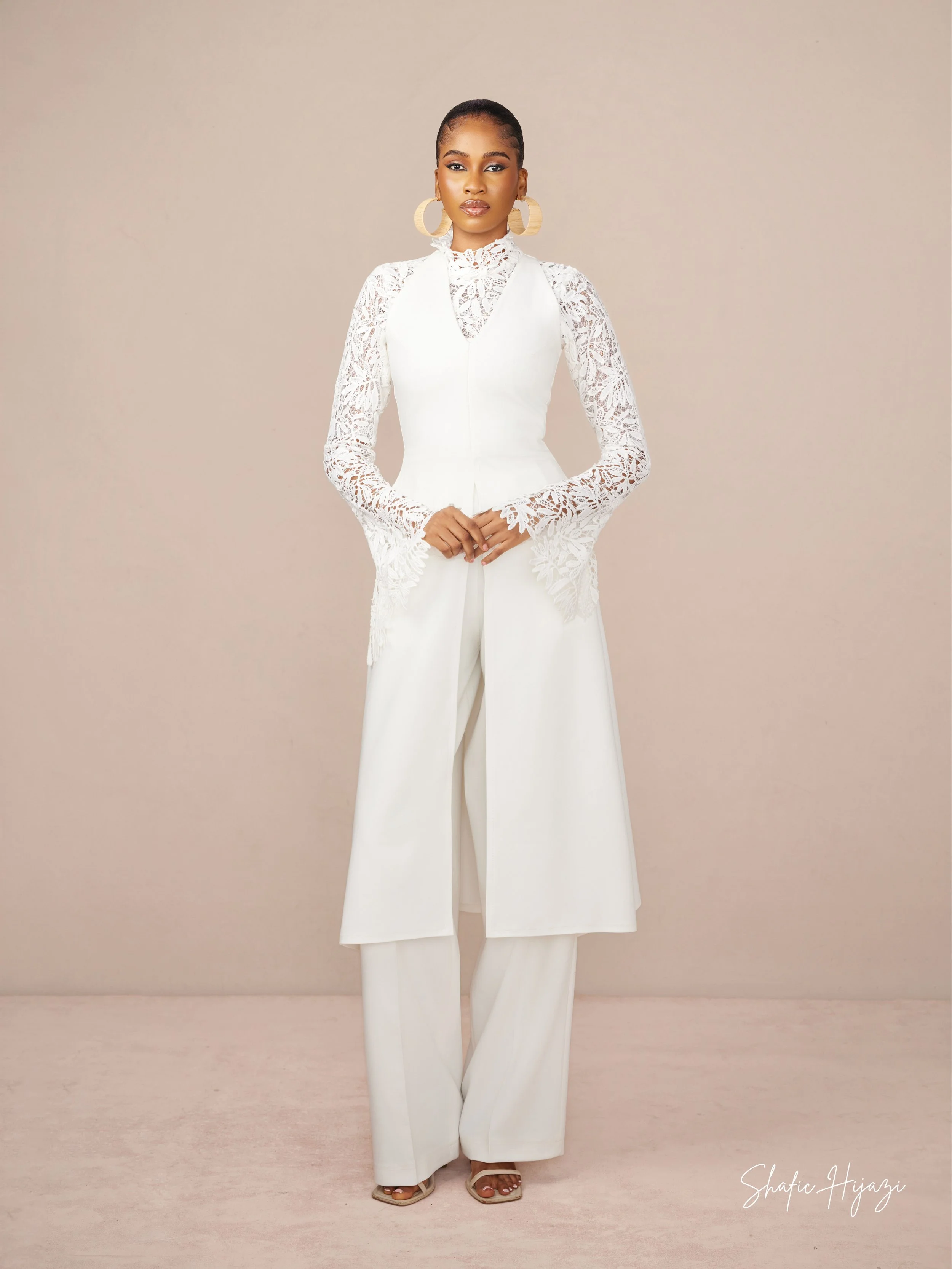 A woman wearing an elegant white outfit with lace sleeves and wide-legged pants, standing against a light pink background.