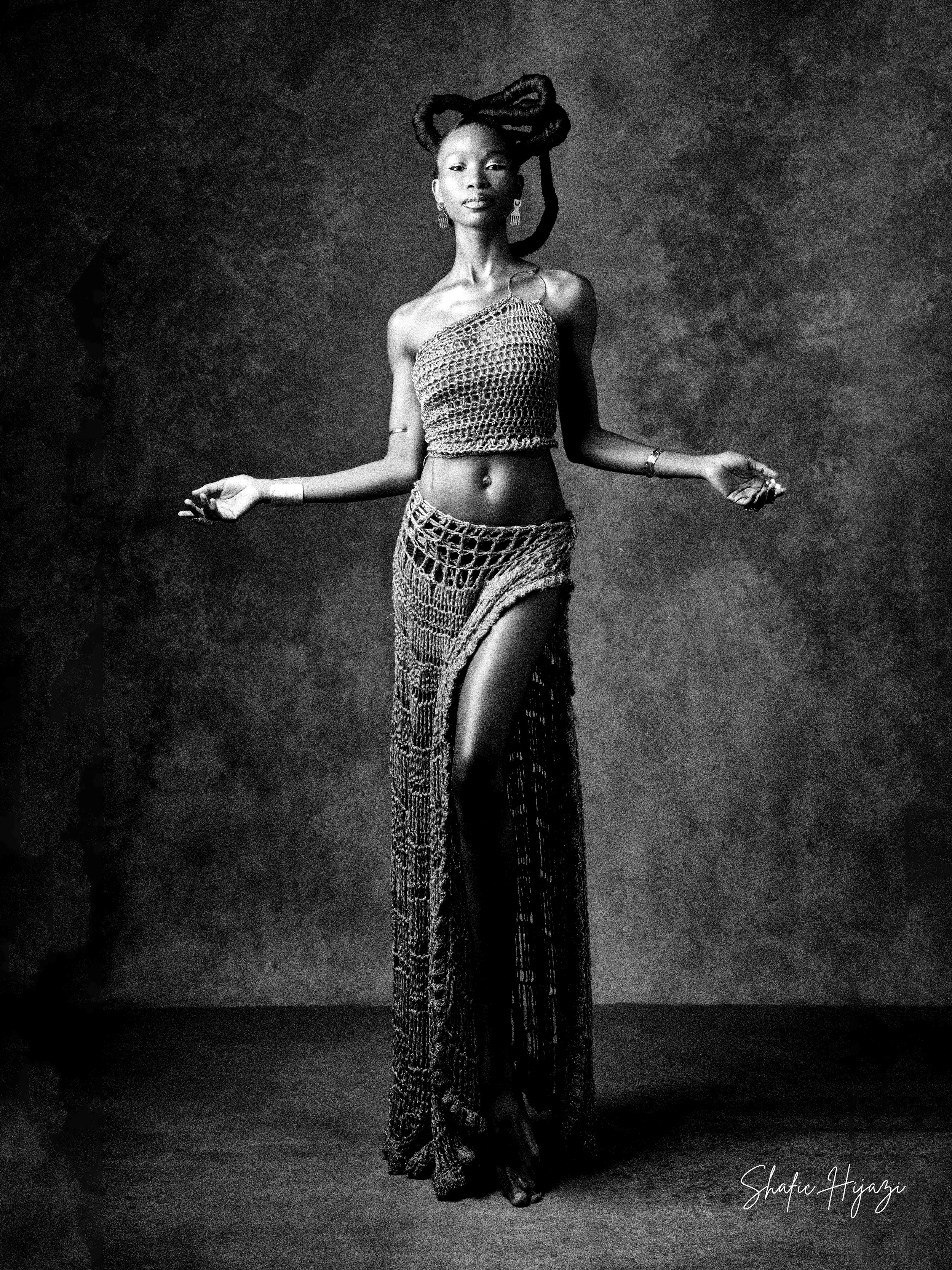 A woman with dreadlocks styled in an intricate updo, wearing a crochet top and long crochet skirt with a high slit, standing against a textured background.