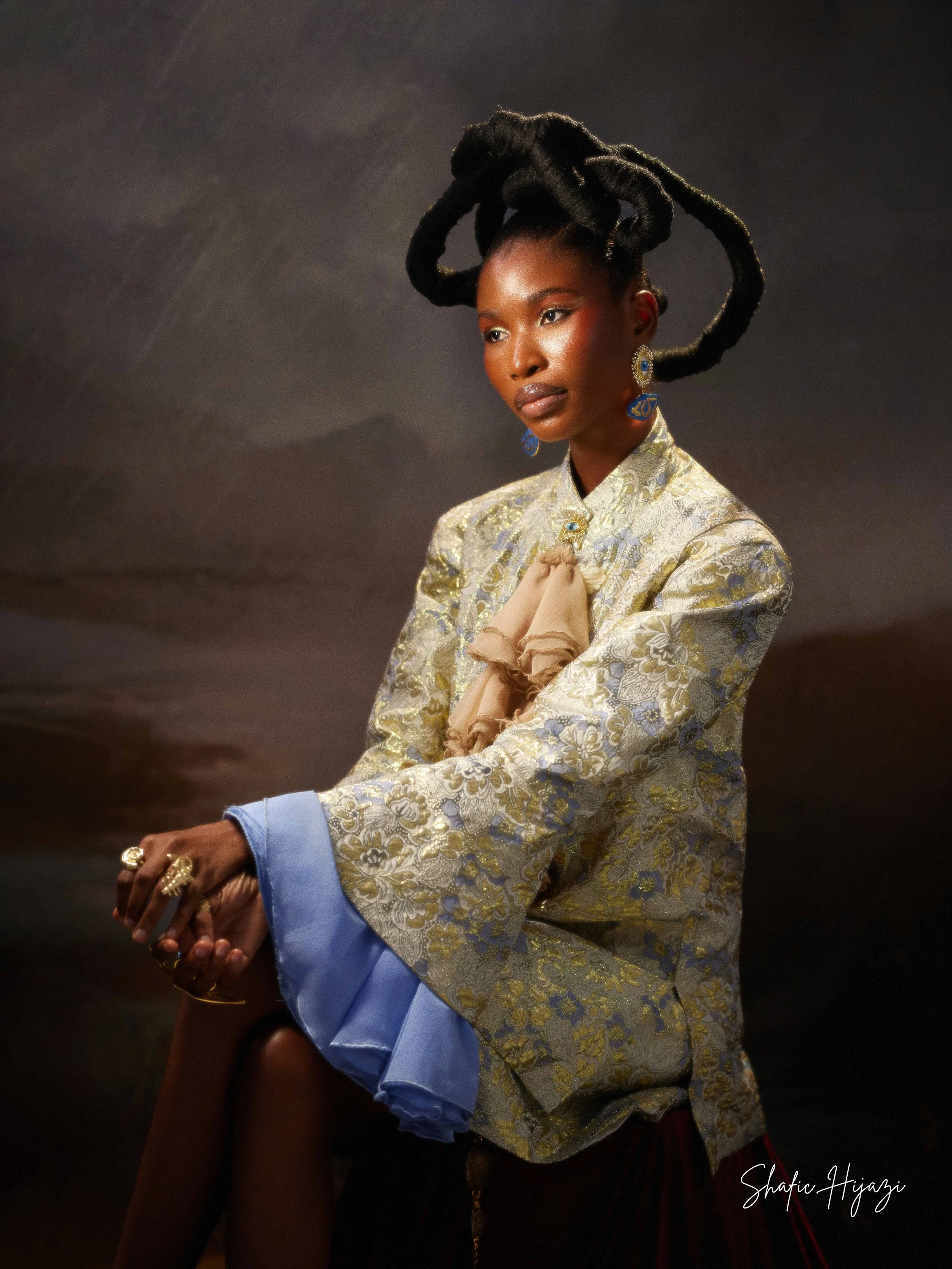 A woman with elaborate, sculptural hair styled in large, coiled twists, wearing a gold and silver brocade jacket with blue accents, statement earrings, rings, and a beige accessory on her chest, against a dark background.