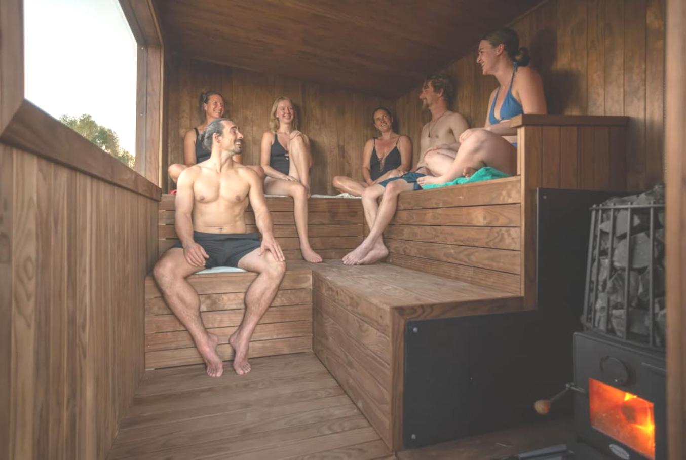 Six people sitting inside a wooden sauna, smiling and enjoying each other’s company.